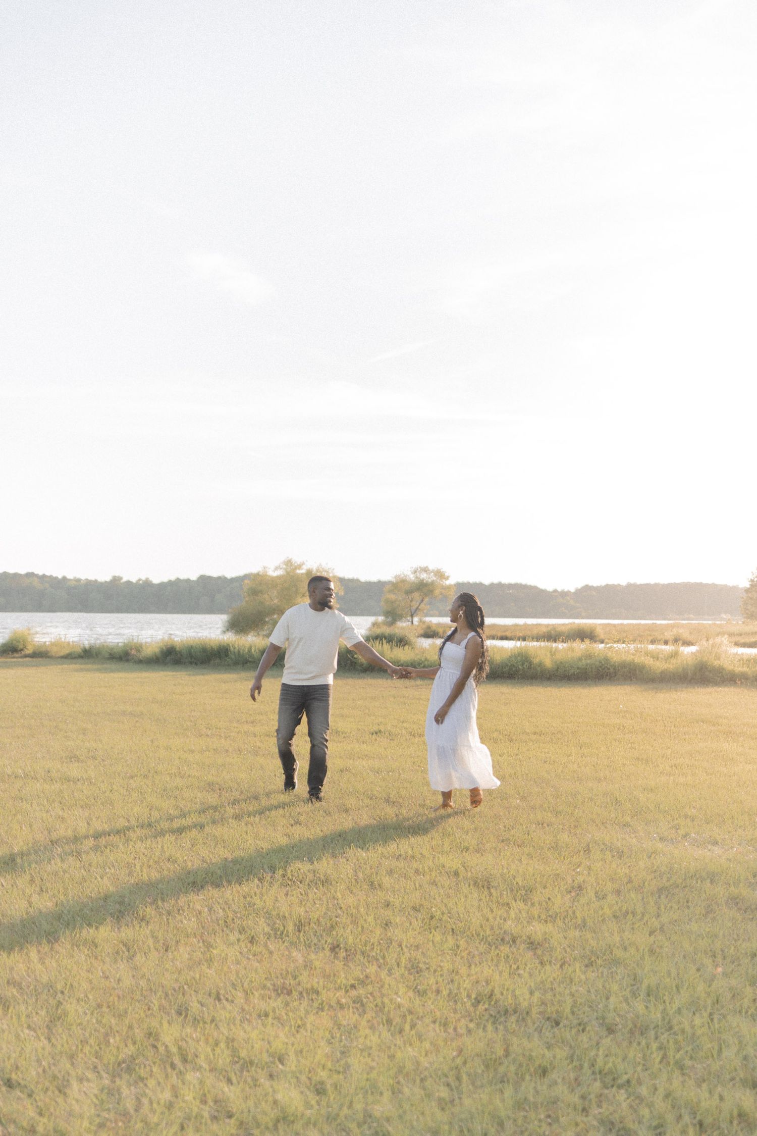 Couple walking hand in hand through a sunlit grassy field near a lake, woman in white dress, man in jeans and white shirt.