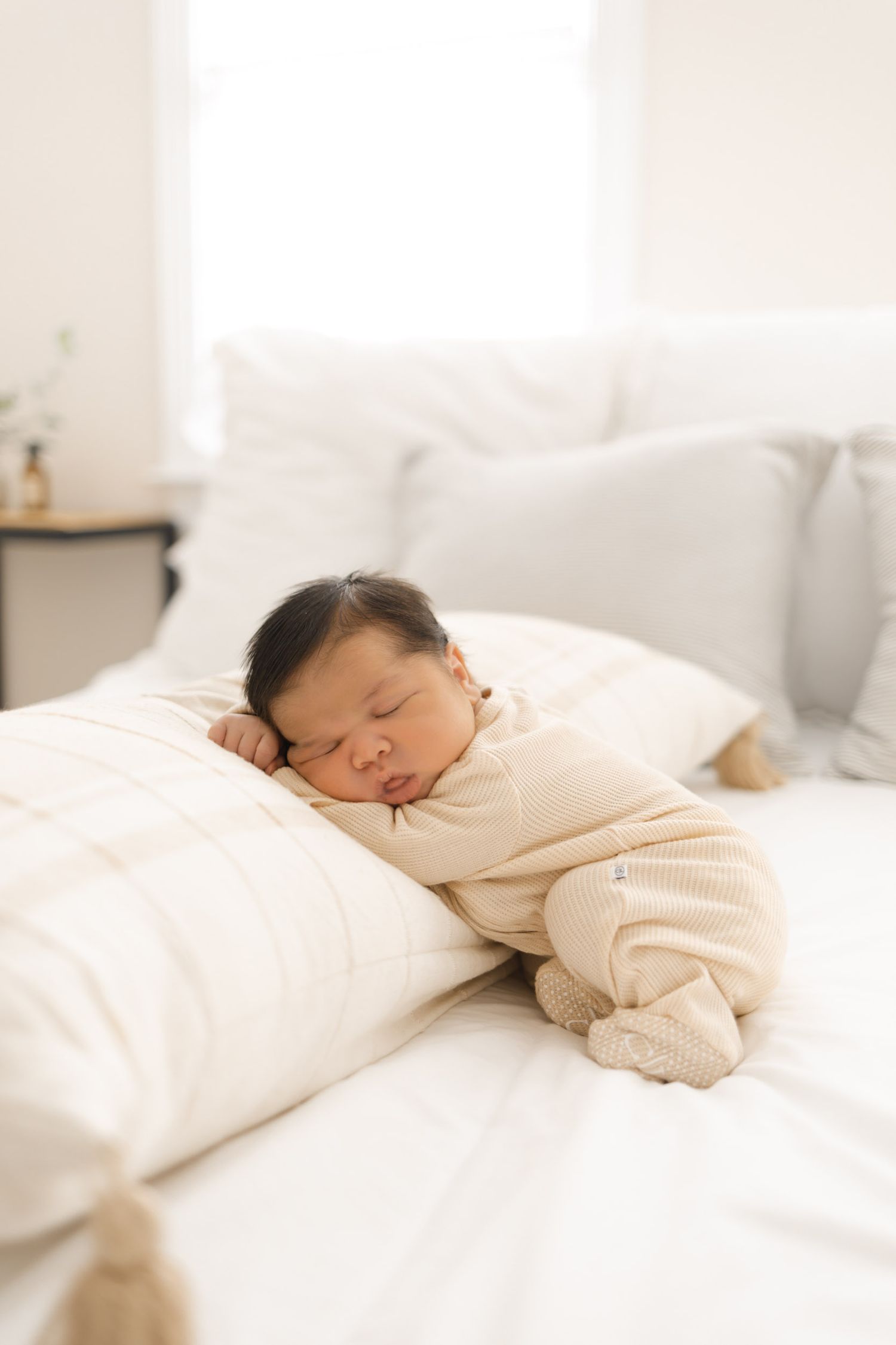 A peaceful newborn baby sleeping on a white bed, dressed in a cozy beige knit outfit, resting against soft pillows.