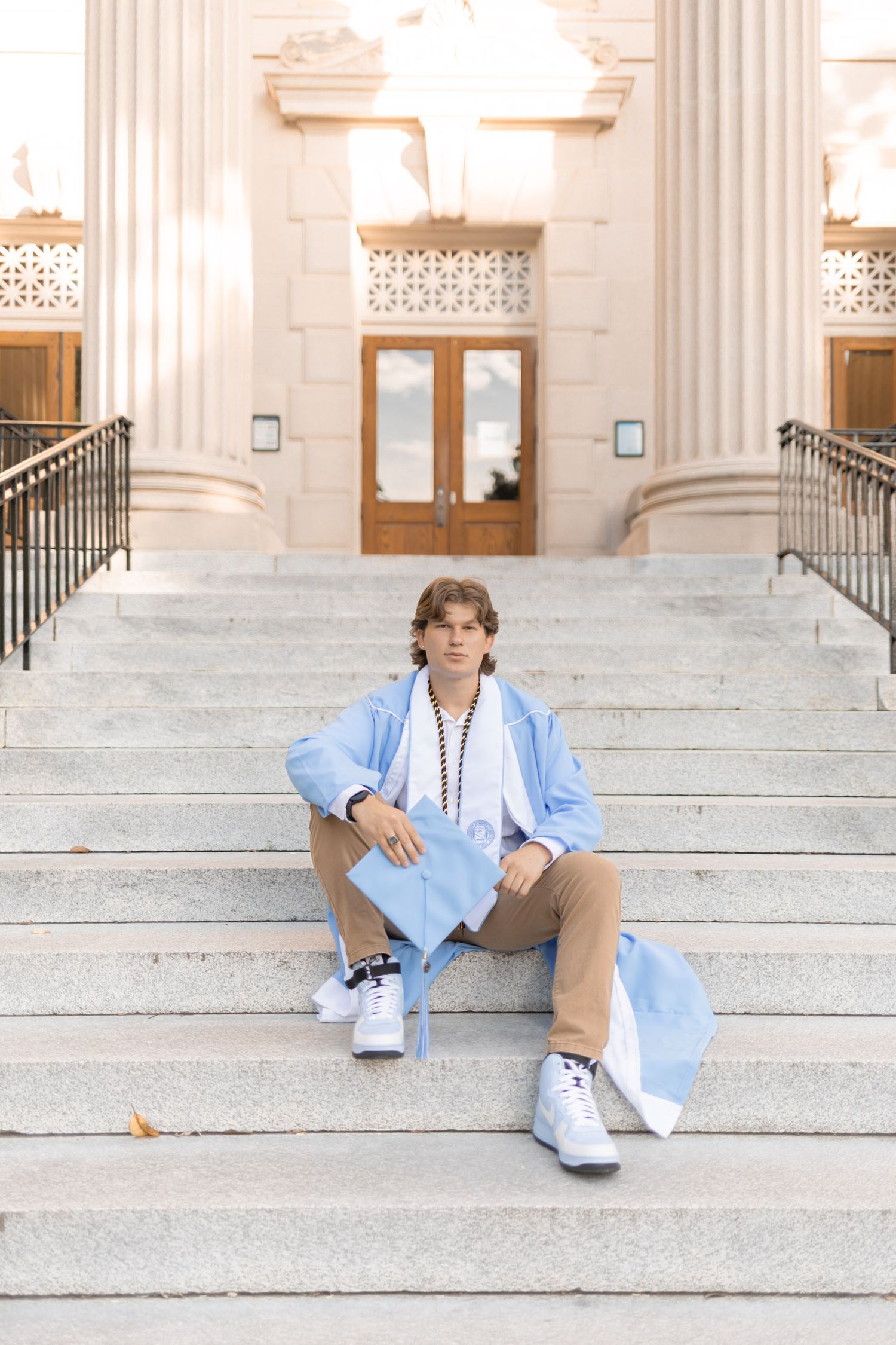 Graduate in light blue gown sits on stone steps outside a grand university building.