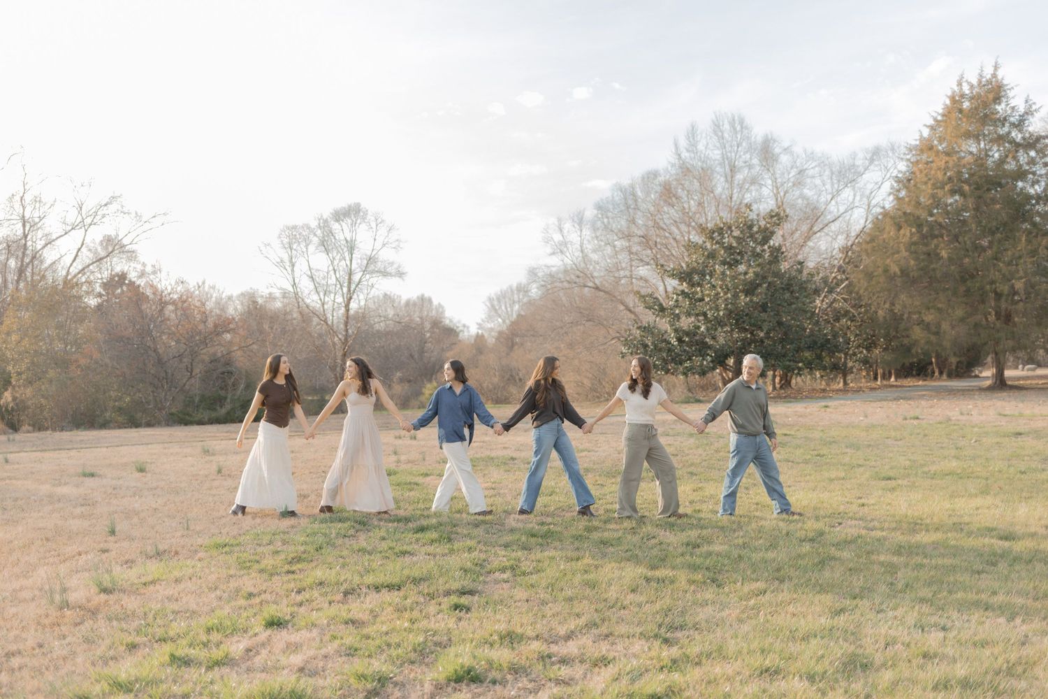 Six people holding hands walking through a sunny open field surrounded by autumn trees.