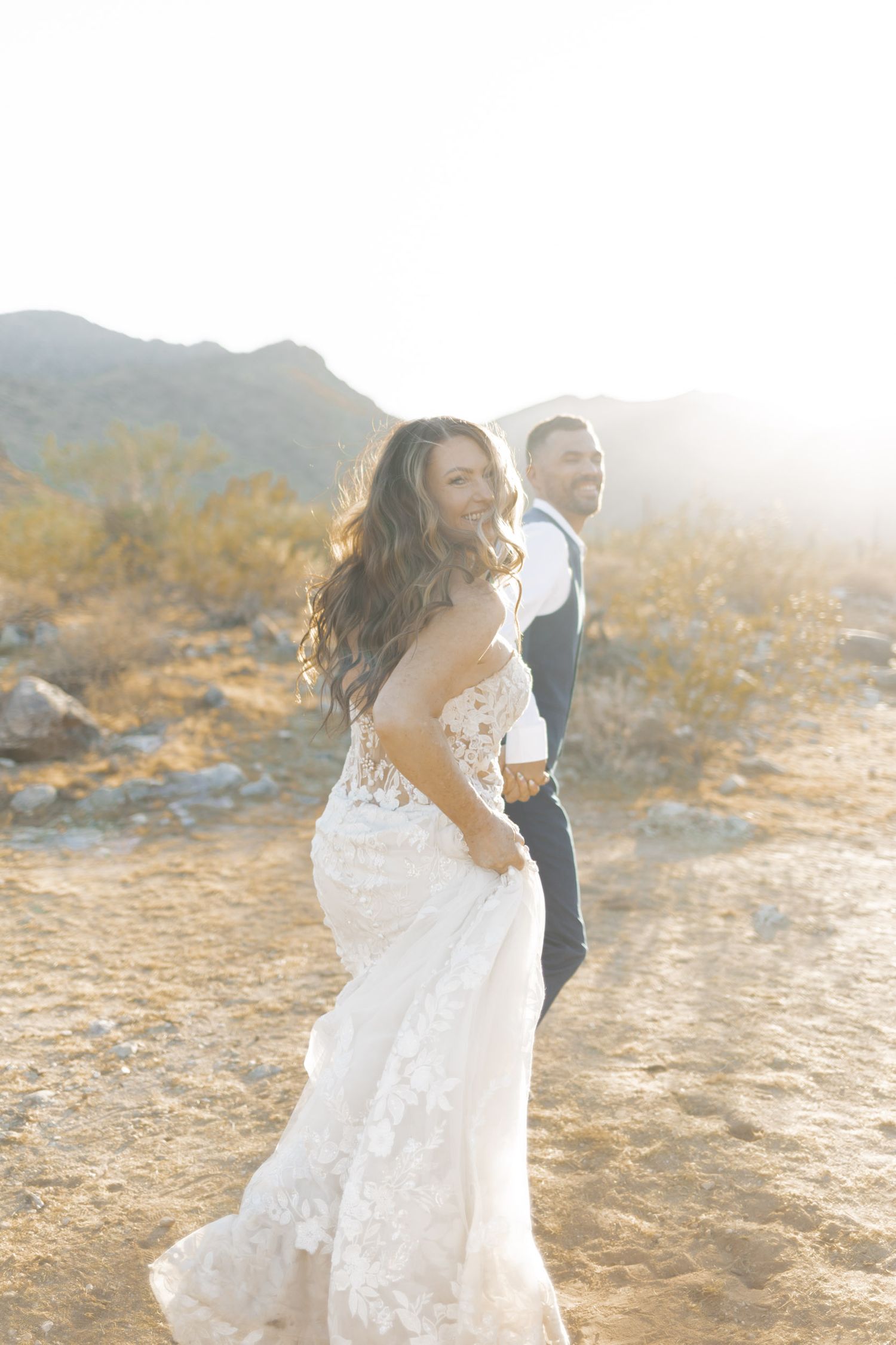 Bride in lace gown smiles over shoulder while groom holds her hand in a sunlit desert landscape.