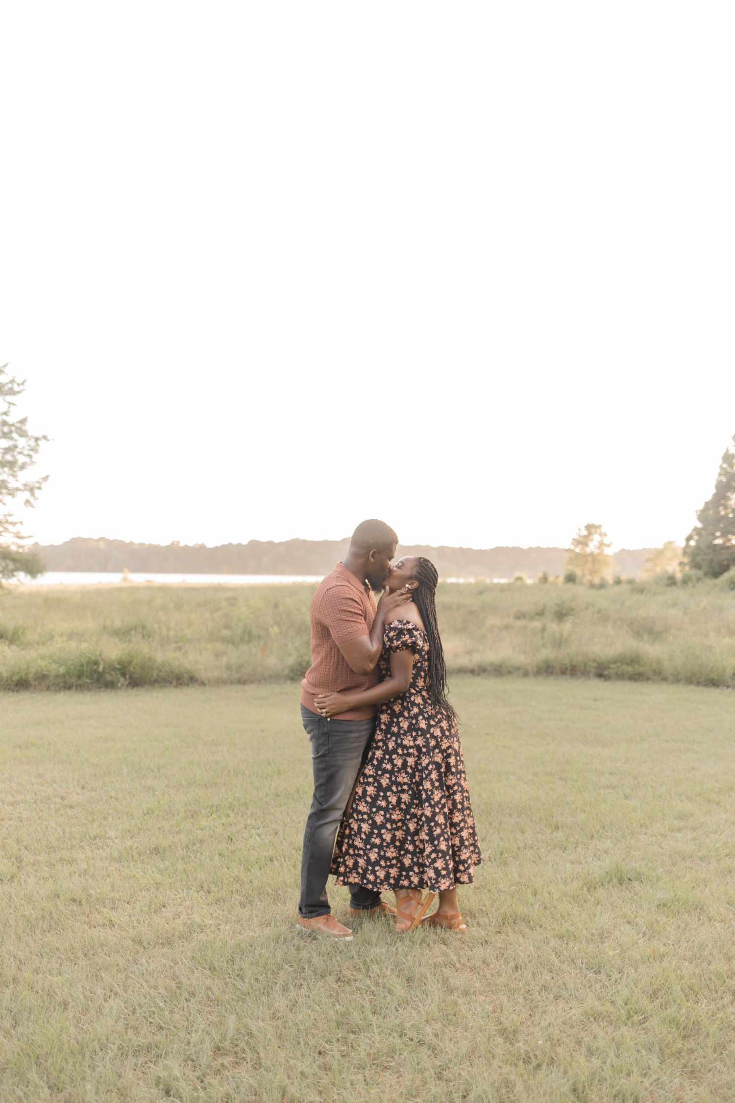 A couple shares a romantic kiss in an open grassy field near a lake during golden hour.