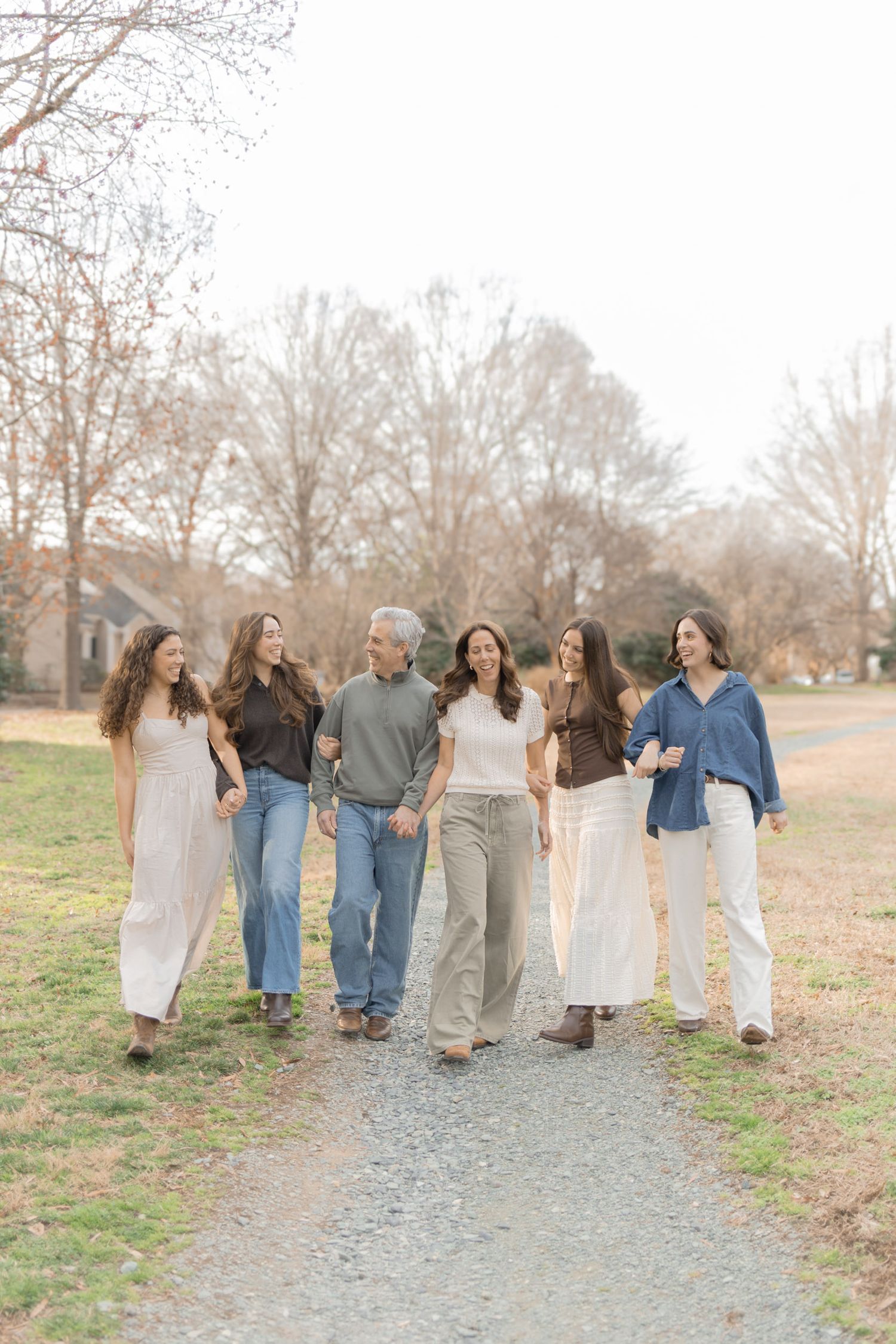 A happy family of six walks together on a gravel path outdoors, dressed in neutral tones, smiling and laughing.