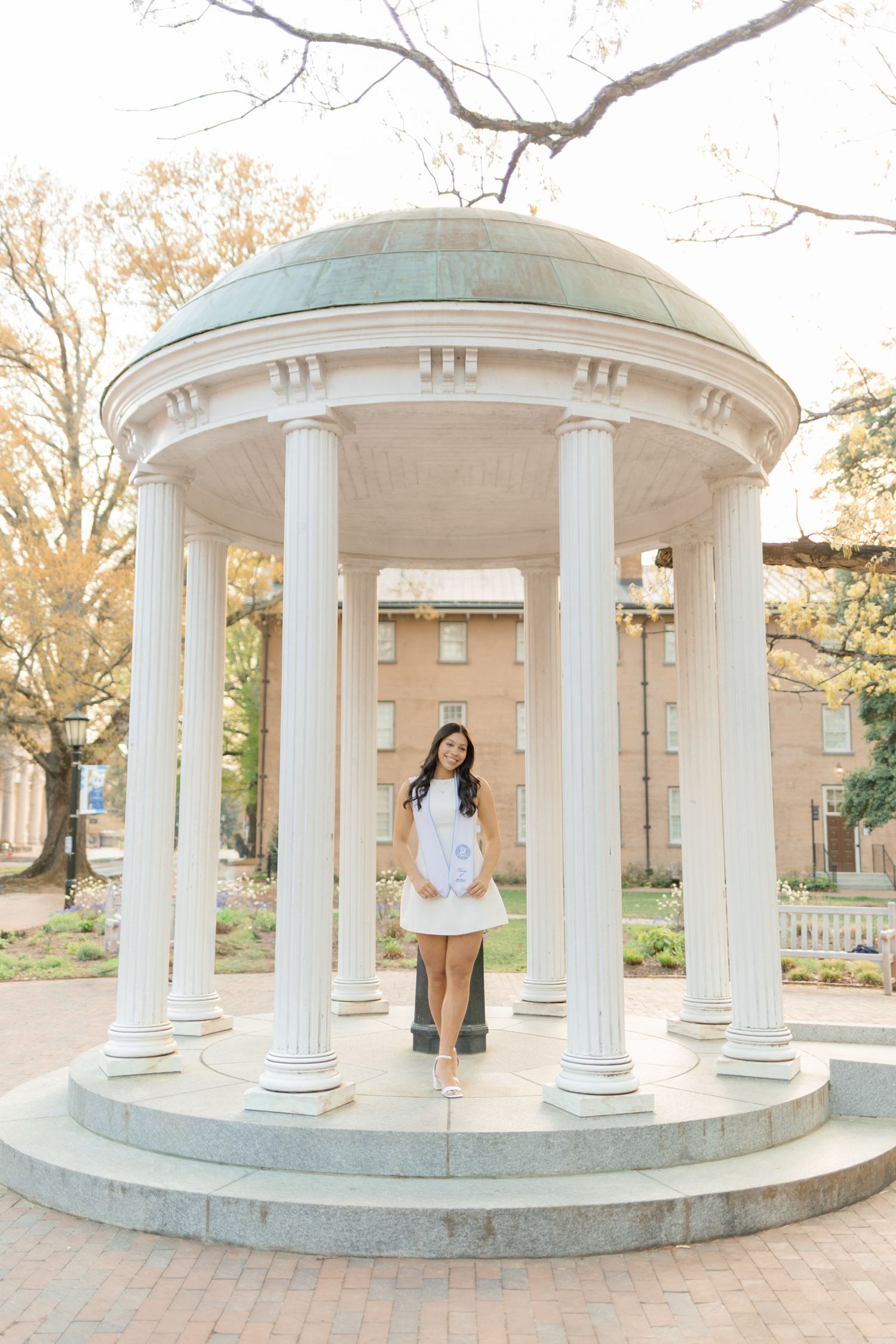 Female graduate in white dress standing inside the iconic Old Well rotunda at UNC.