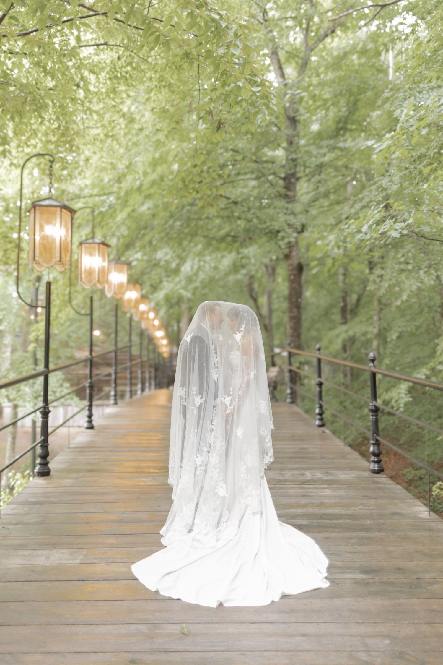 Bride in white gown and flowing veil stands on wooden bridge surrounded by lush green trees and glowing lanterns.