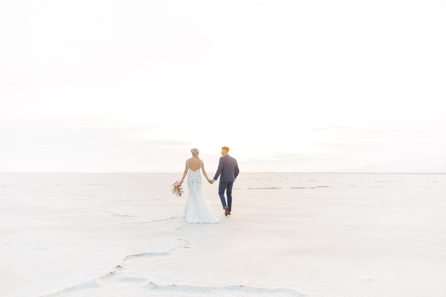 A couple walks hand in hand across a bright white salt flat landscape with mountains in the distance.