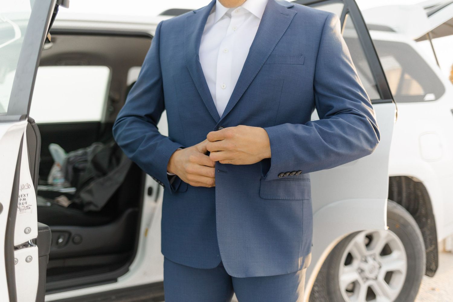 A groom in a navy suit adjusts his tie while standing next to a white car.