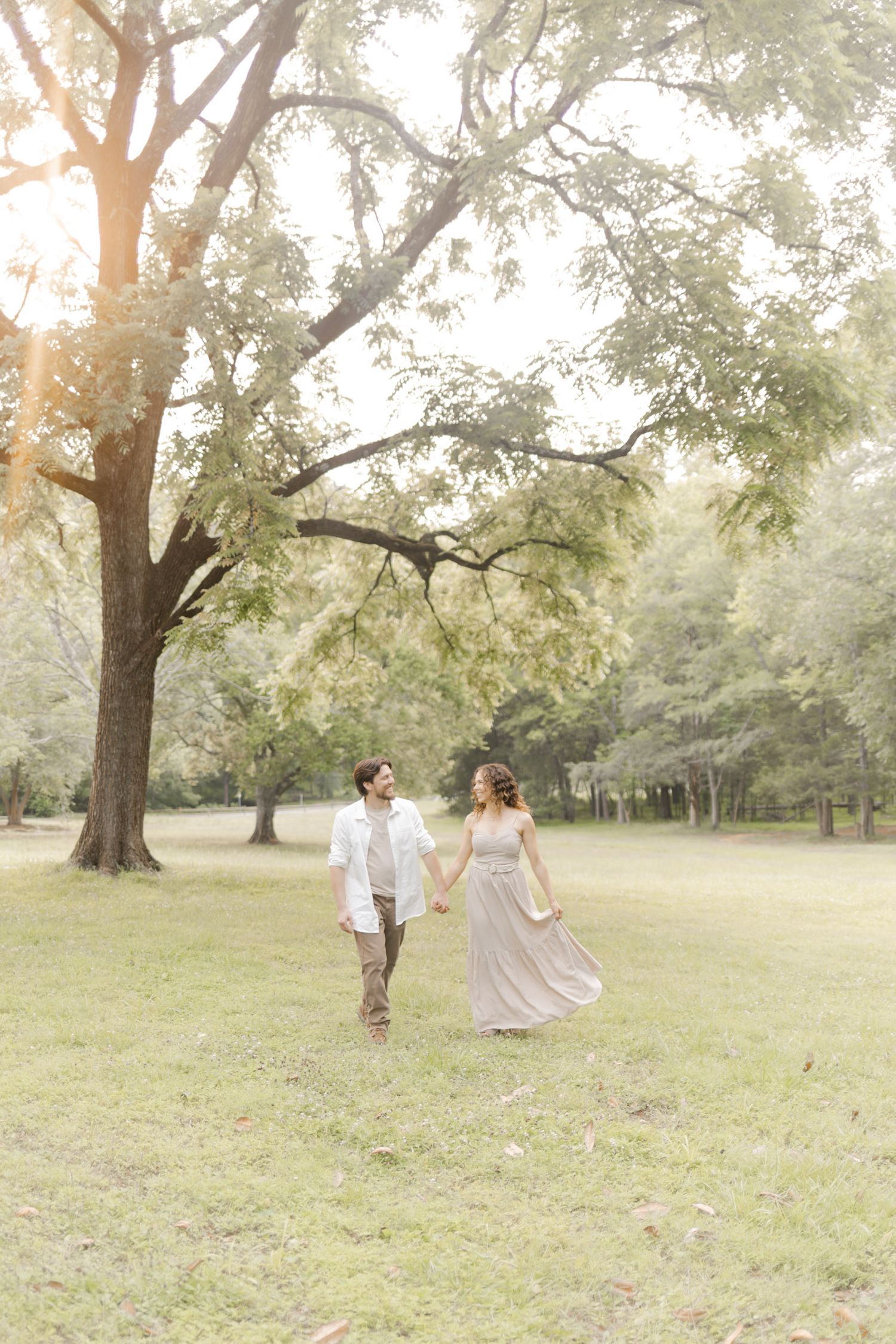 Two people in light colored clothing walk through a sunlit park with large trees overhead during photoshoot with Kinsley Nicole Photography.