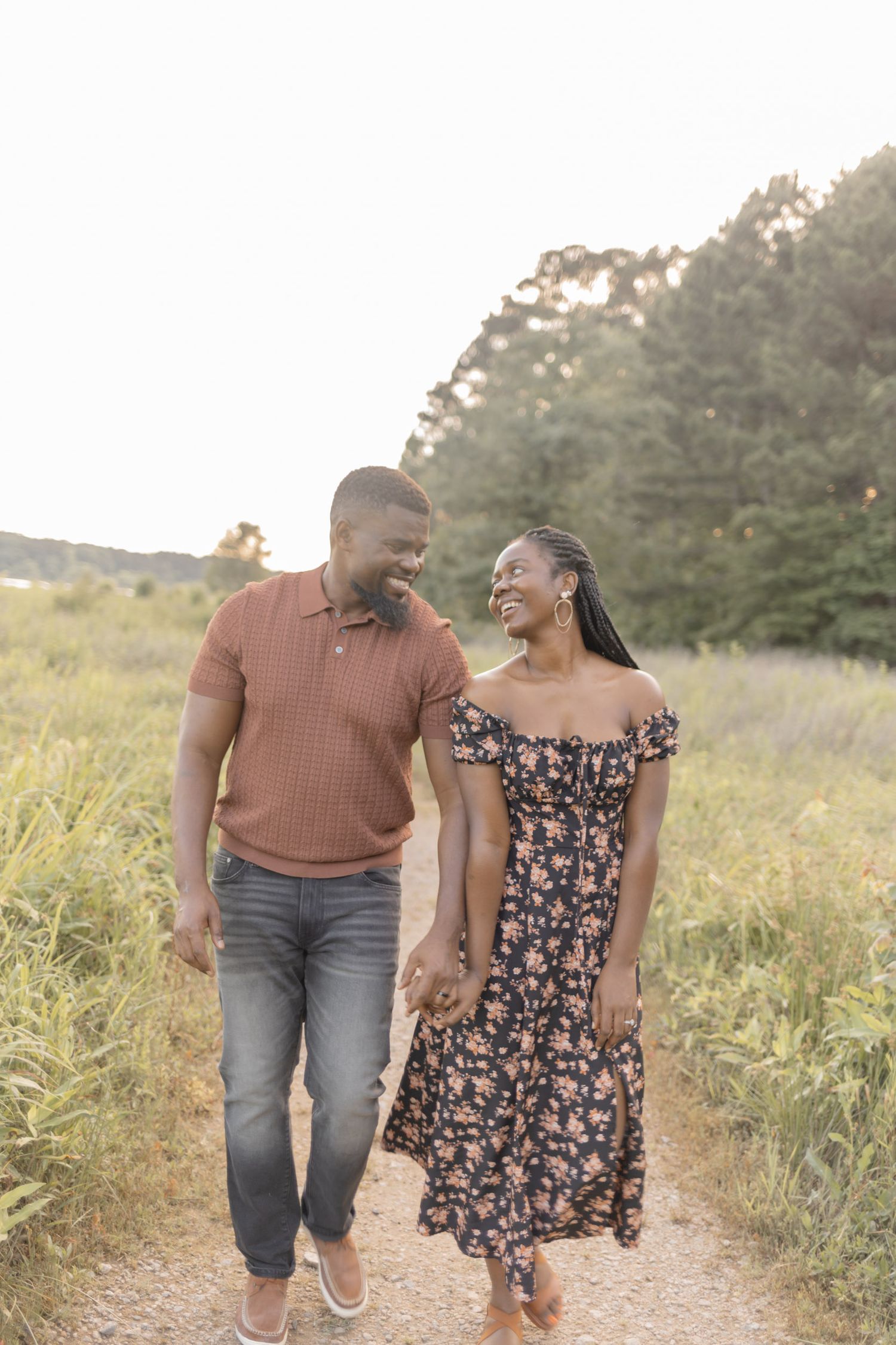 A joyful couple walks hand-in-hand along a sunlit path surrounded by lush greenery, smiling at each other.