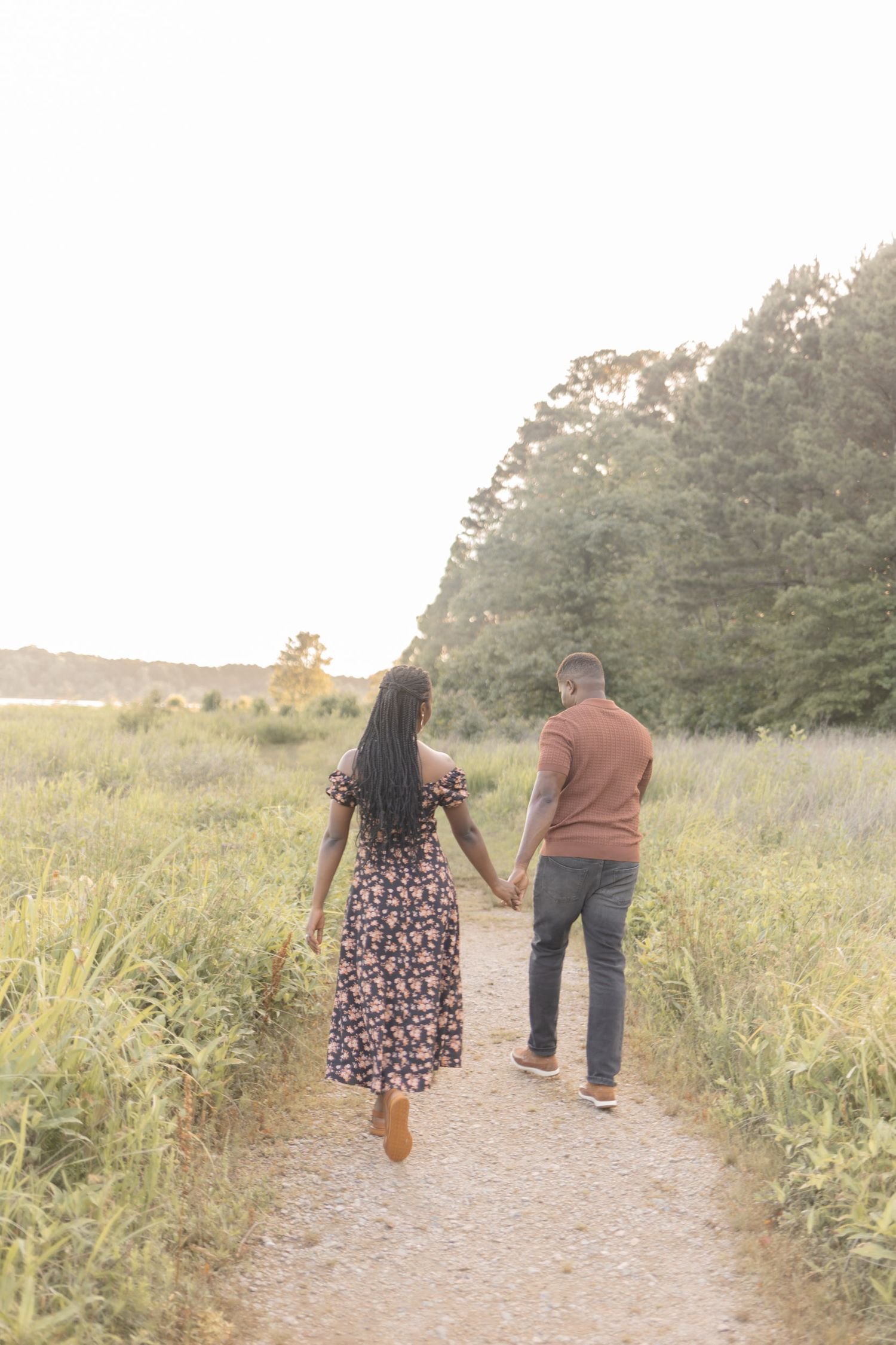 Couple holding hands walking down a scenic path surrounded by lush green fields and trees at golden hour.