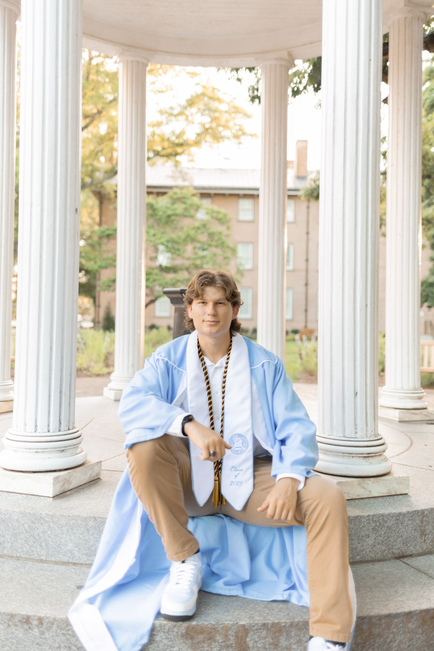 Young man in light blue graduation gown and stole sitting at the Old Well colonnade at UNC Chapel Hill.
