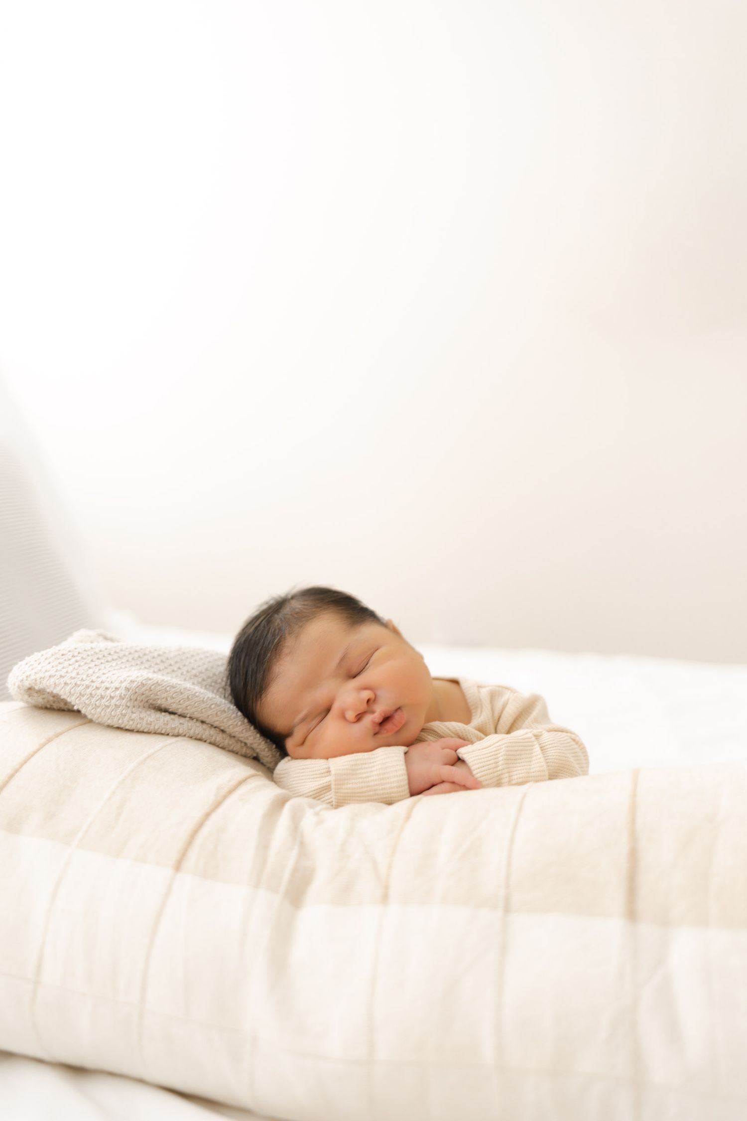 Newborn baby peacefully sleeping on white quilted blanket in bright airy nursery setting.