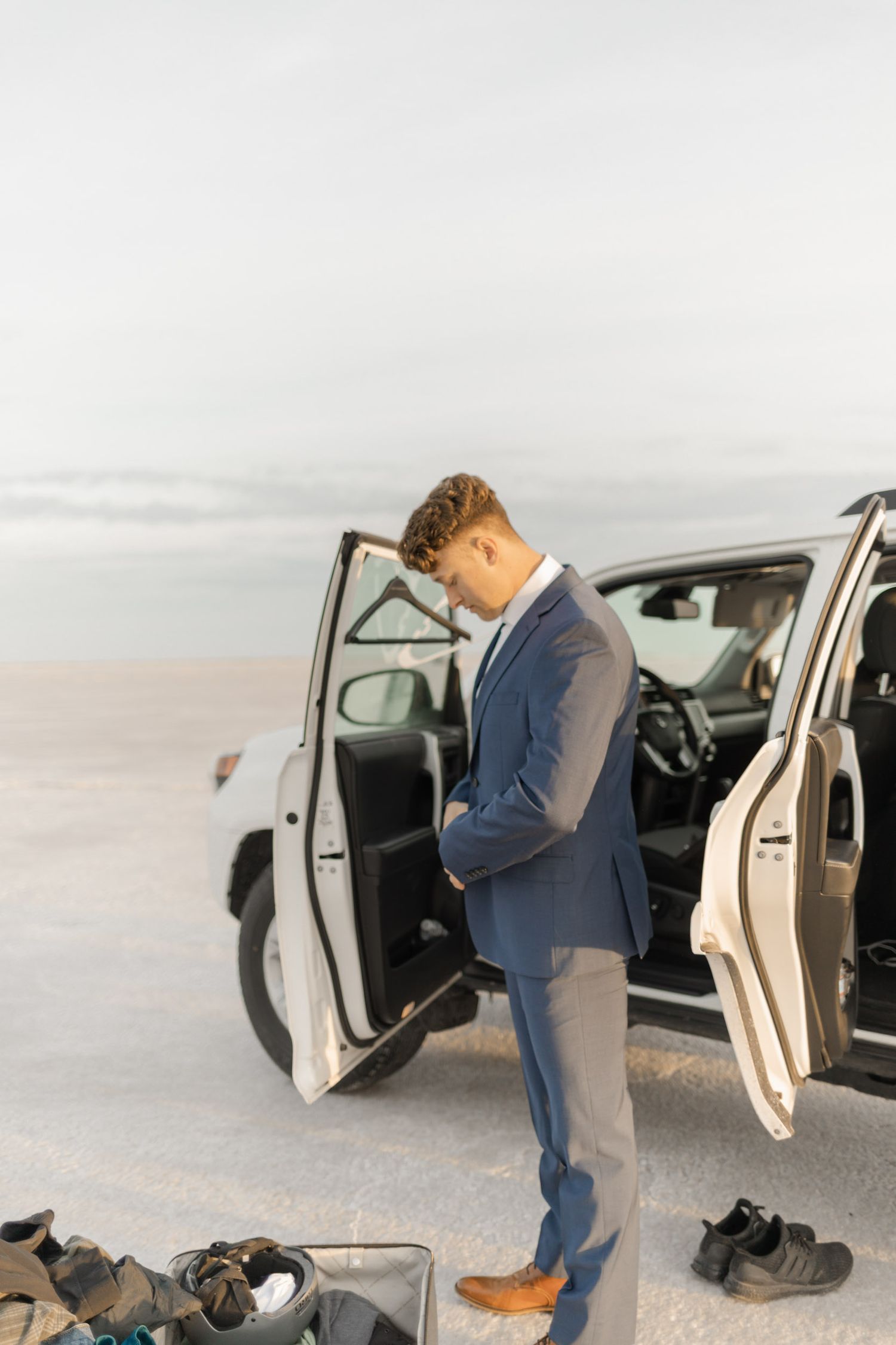 A person in a suit bends down to inspect the interior of a white car door on a salt flat.