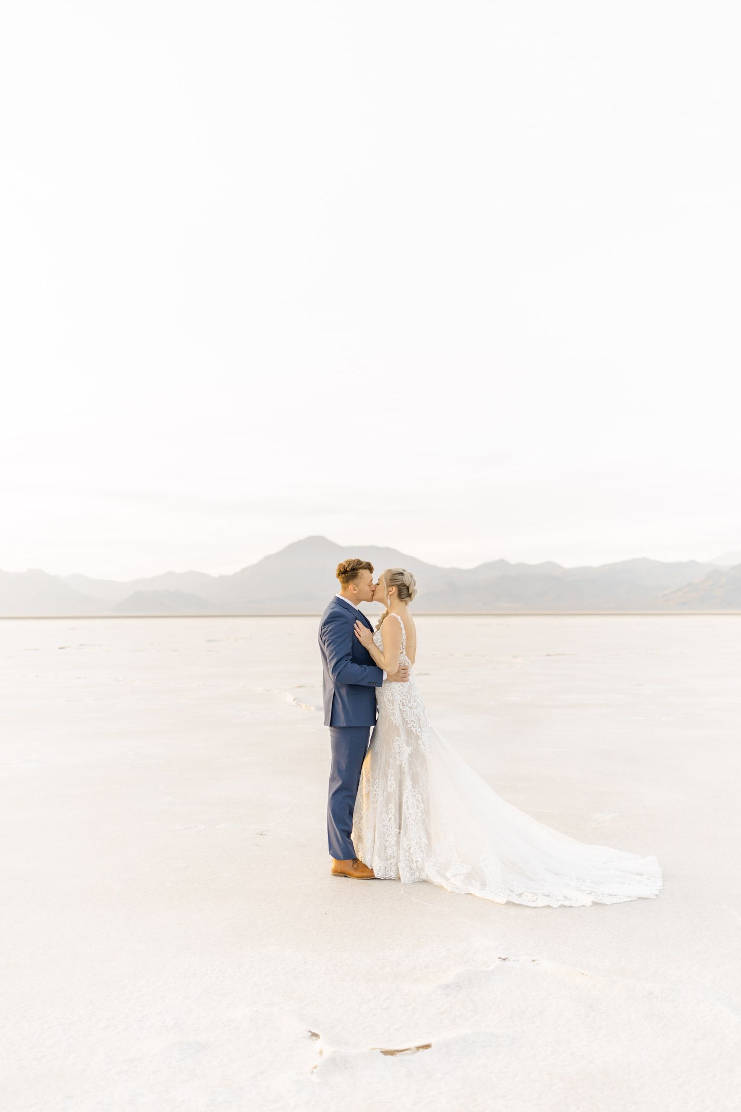 A couple shares an intimate moment on a white sandy beach with mountains in the distance during their wedding day.