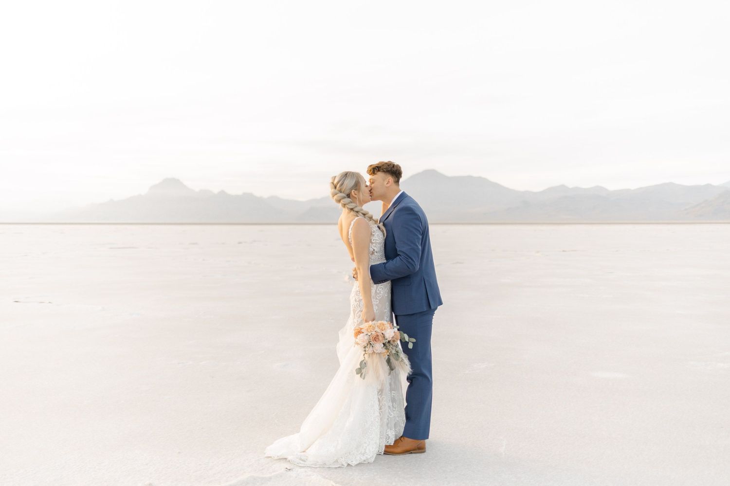 Wedding couple embracing on pristine white salt flats with distant mountain range at sunset.