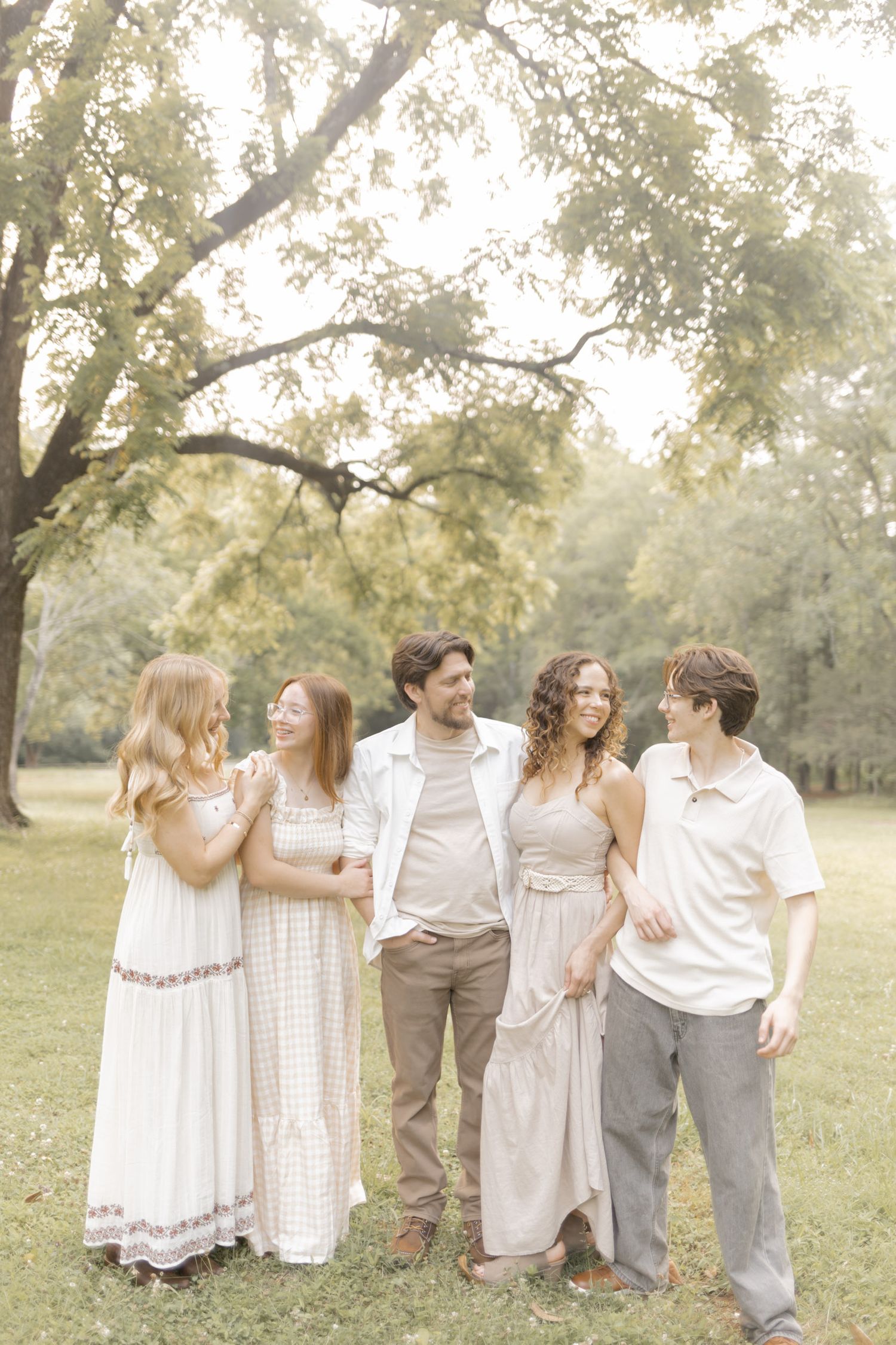 A joyful family of five poses together in a lush green park, wearing coordinated neutral and white outfits.