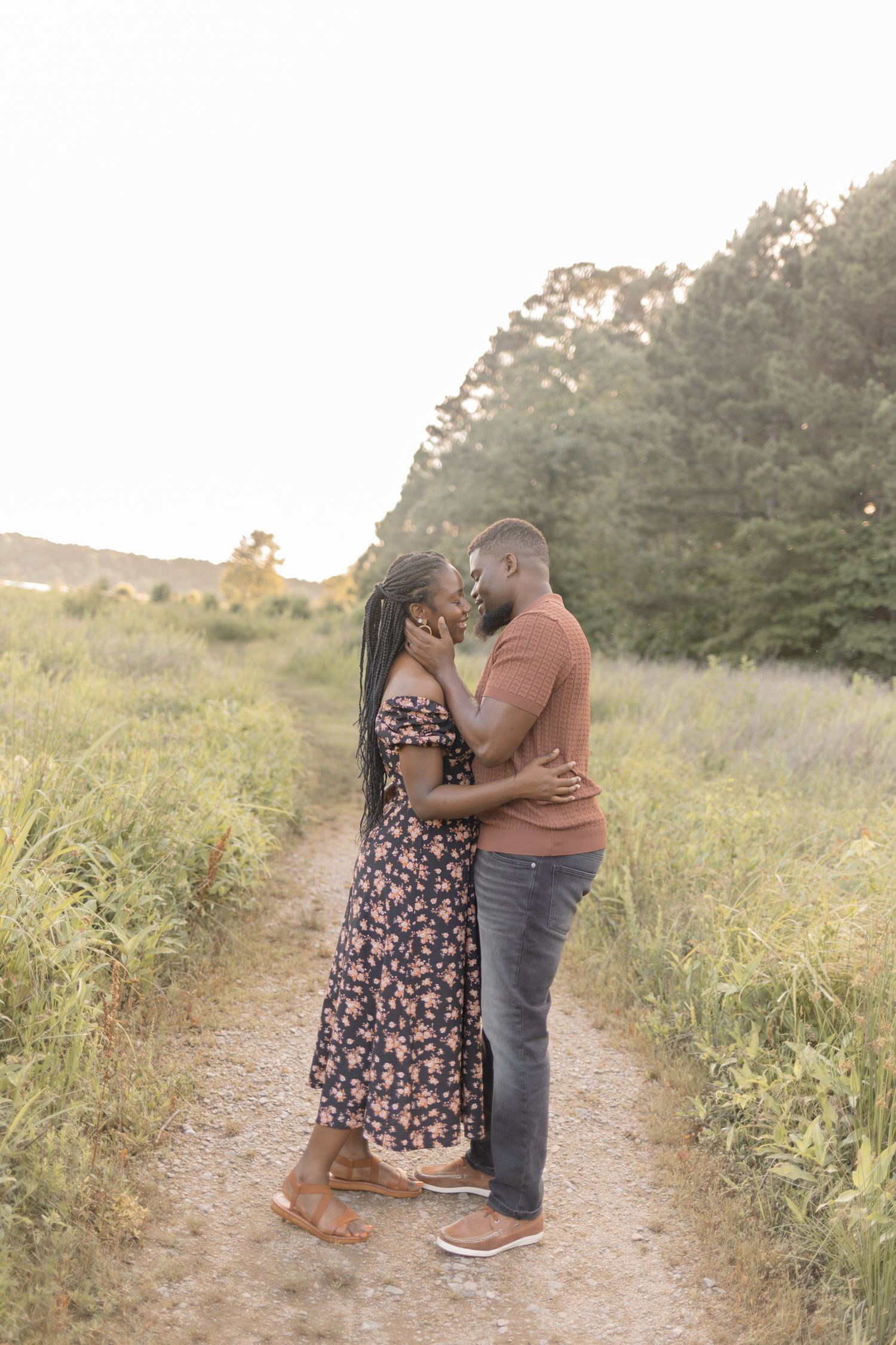 Couple sharing a romantic kiss on a scenic dirt path surrounded by lush greenery at golden hour.