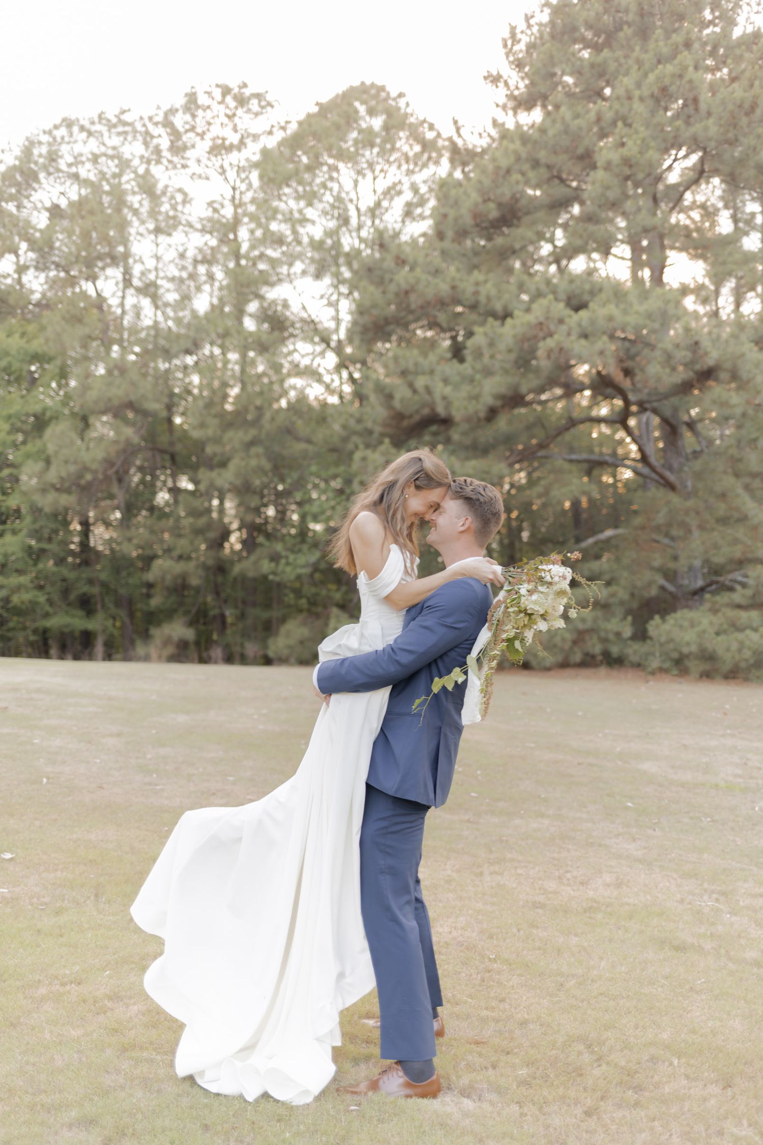 Groom in blue suit lifts bride in white gown, sharing a romantic moment outdoors with pine trees in background.