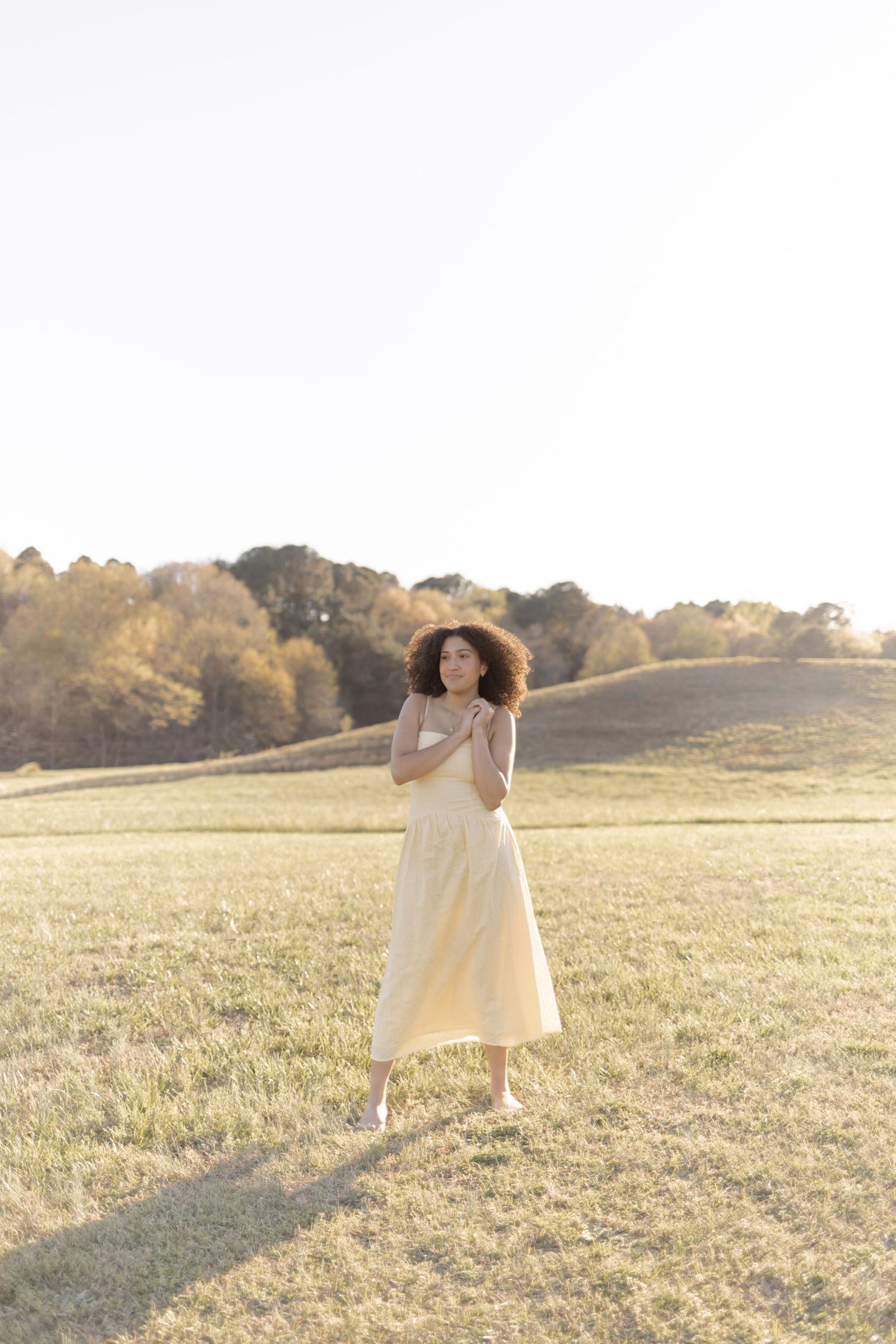 Woman in cream midi dress standing in golden sunlit field with rolling hills and autumn trees in background.