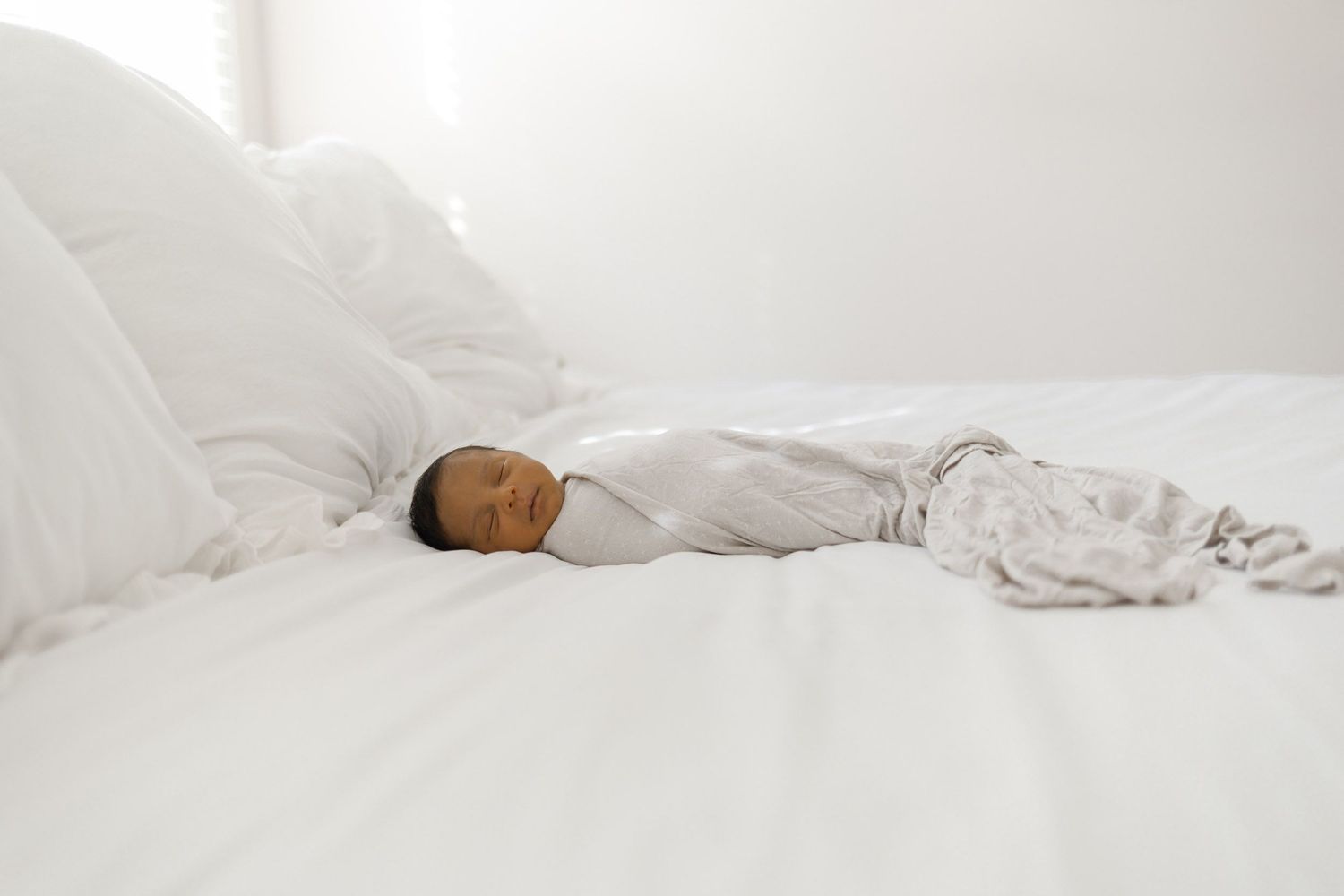 Newborn baby swaddled in gray muslin blanket sleeping on a white bed in bright light.