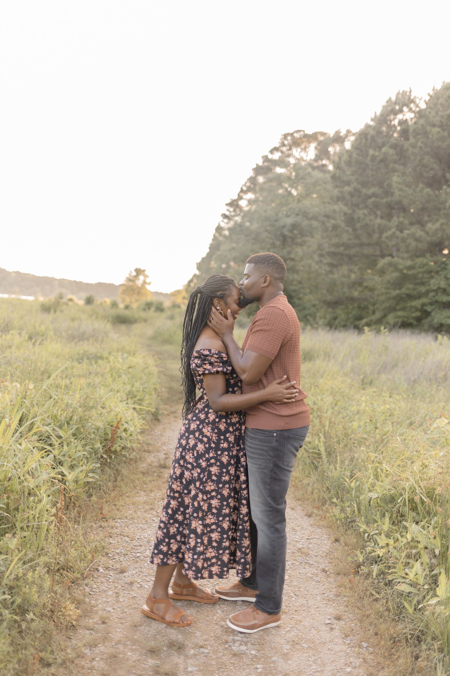 A romantic couple shares a kiss on a scenic dirt path surrounded by lush greenery at golden hour.