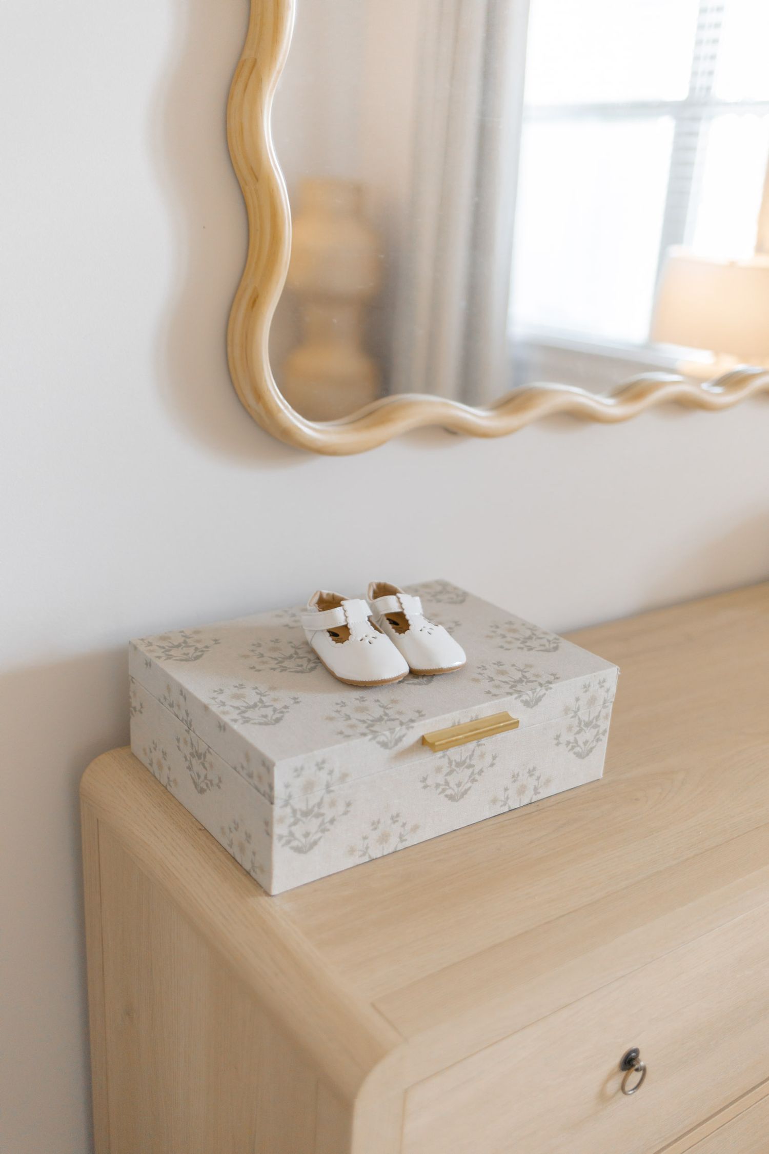 Tiny white baby Mary Jane shoes rest on a floral keepsake box atop a light wood dresser beneath a gold wavy mirror.