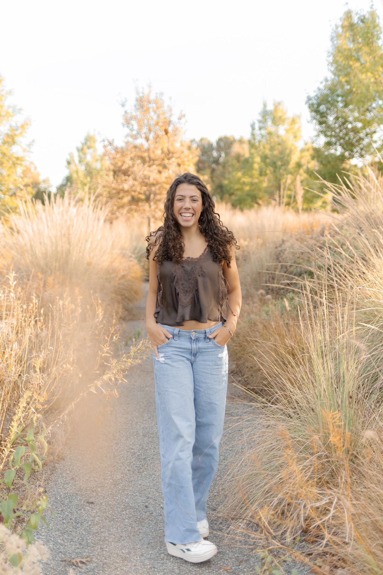 Young woman with curly hair smiling on a nature path, wearing brown top and light blue jeans.