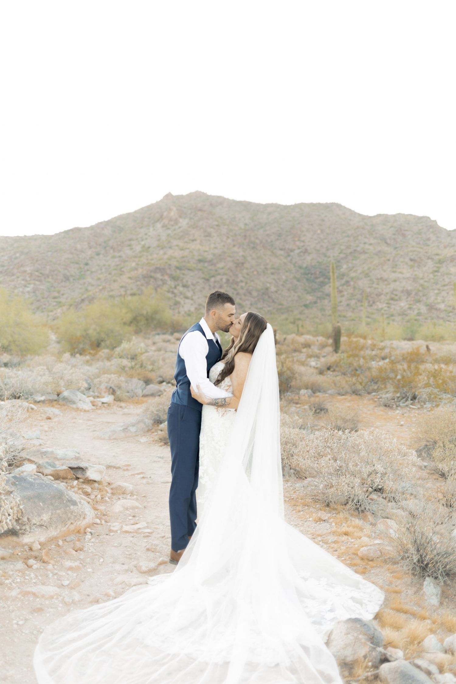 A bride and groom share a romantic kiss in the Arizona desert landscape, with mountains and cacti in the background.