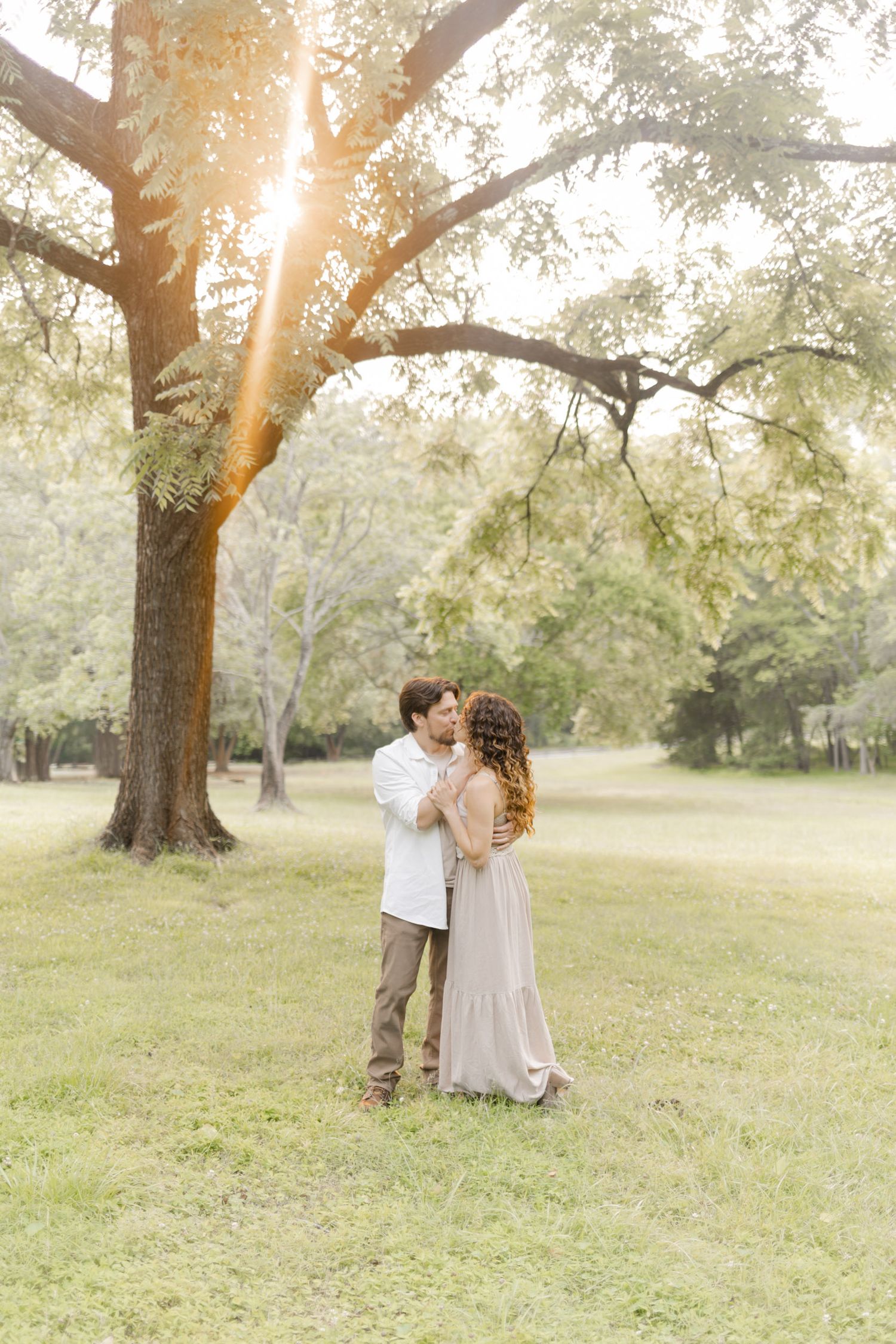 Couple shares intimate moments in a dreamy sunlit park setting wearing neutral toned clothing while doing a photoshoot.