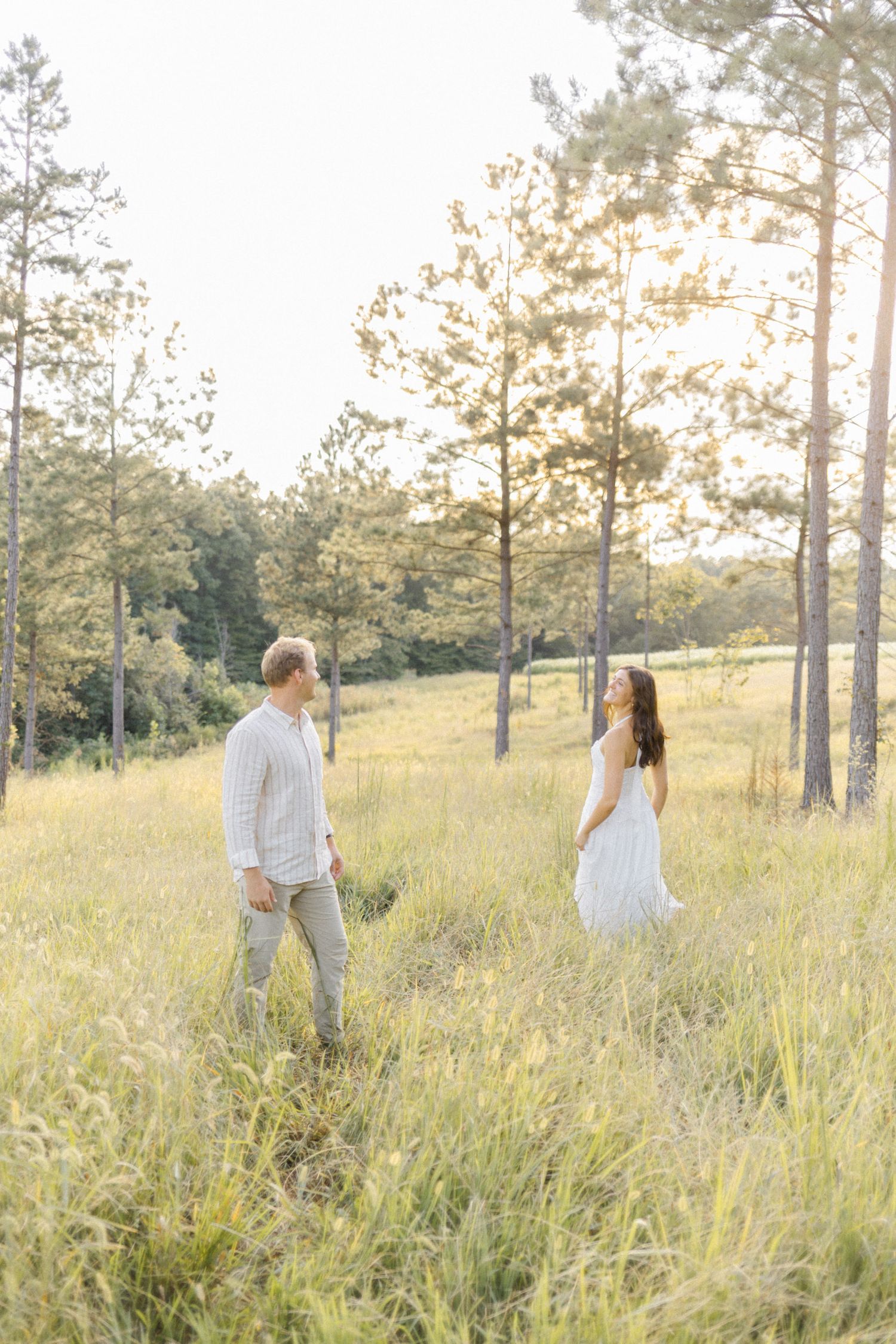 Couple in white outfits walking through a sunlit meadow with tall grass and pine trees at golden hour during lifestyle portrait photoshoot.