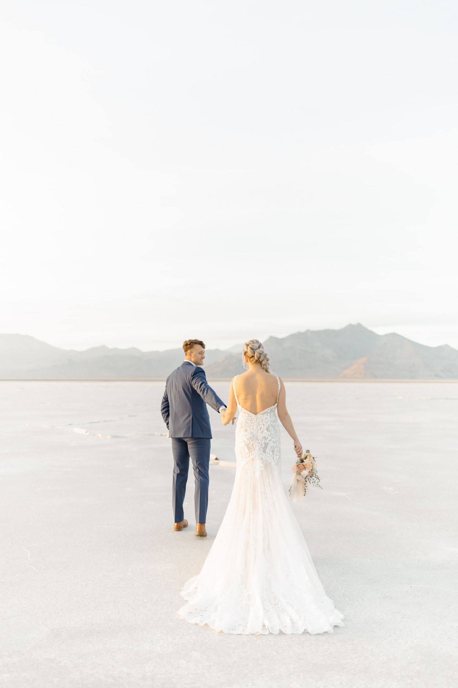 A couple in wedding attire walks hand in hand across the ethereal white salt flats with mountains in the distance.