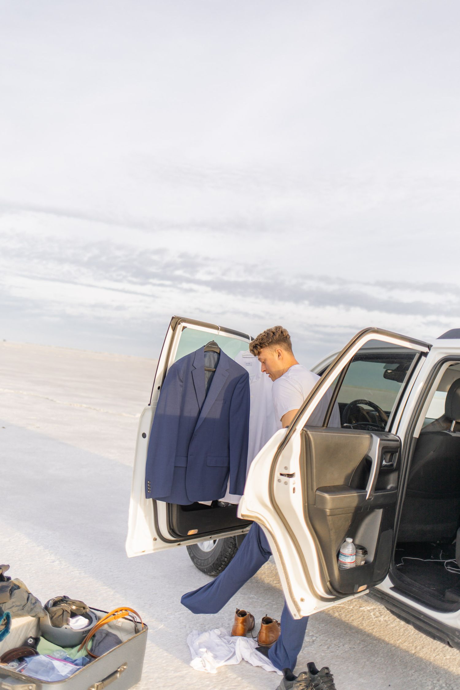 A car door is open on a beach with a navy suit jacket hanging and clothing items scattered on the ground.