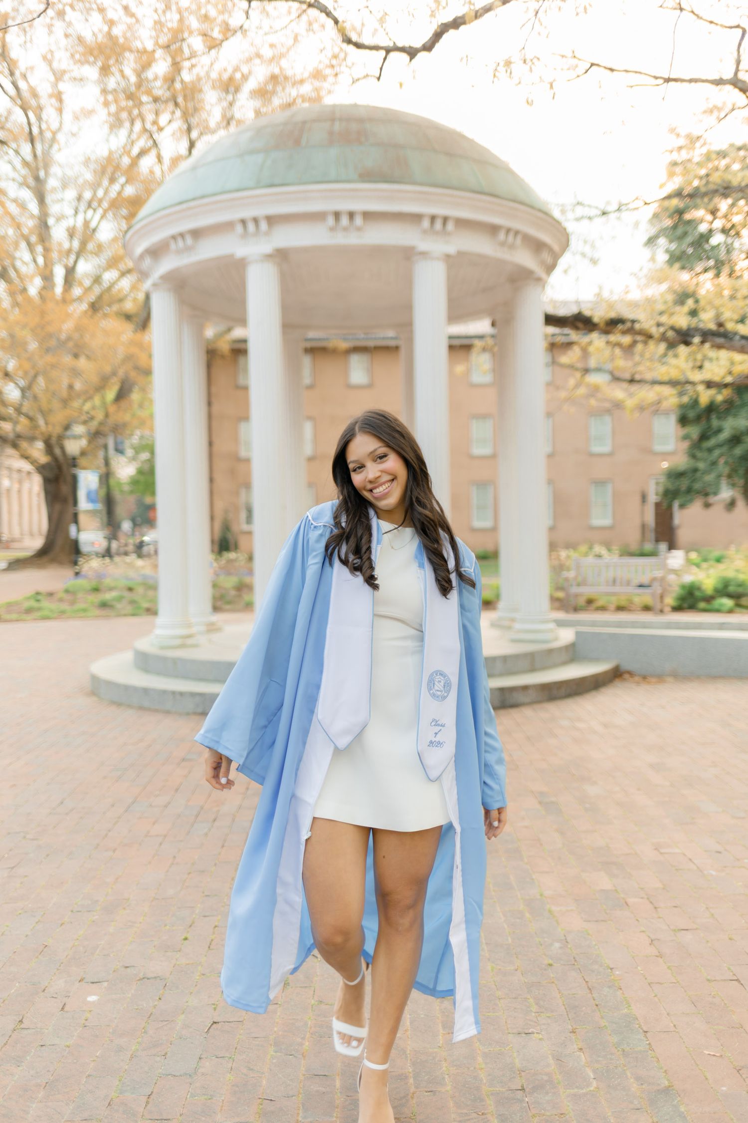 Smiling graduate in light blue cap and gown poses in front of iconic Old Well at UNC Chapel Hill campus.