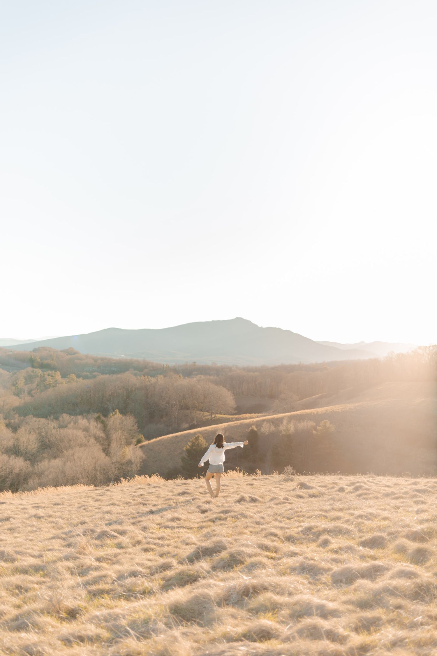 Woman in white dress running through golden meadow with mountain landscape and warm sunset glow.