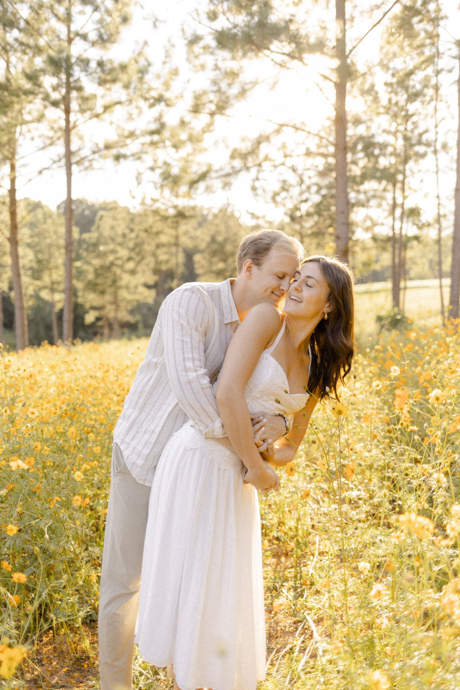 Romantic couple embracing in a field of yellow flowers with pine trees and warm sunset light for engagement photoshoot in Chapel Hill, NC.