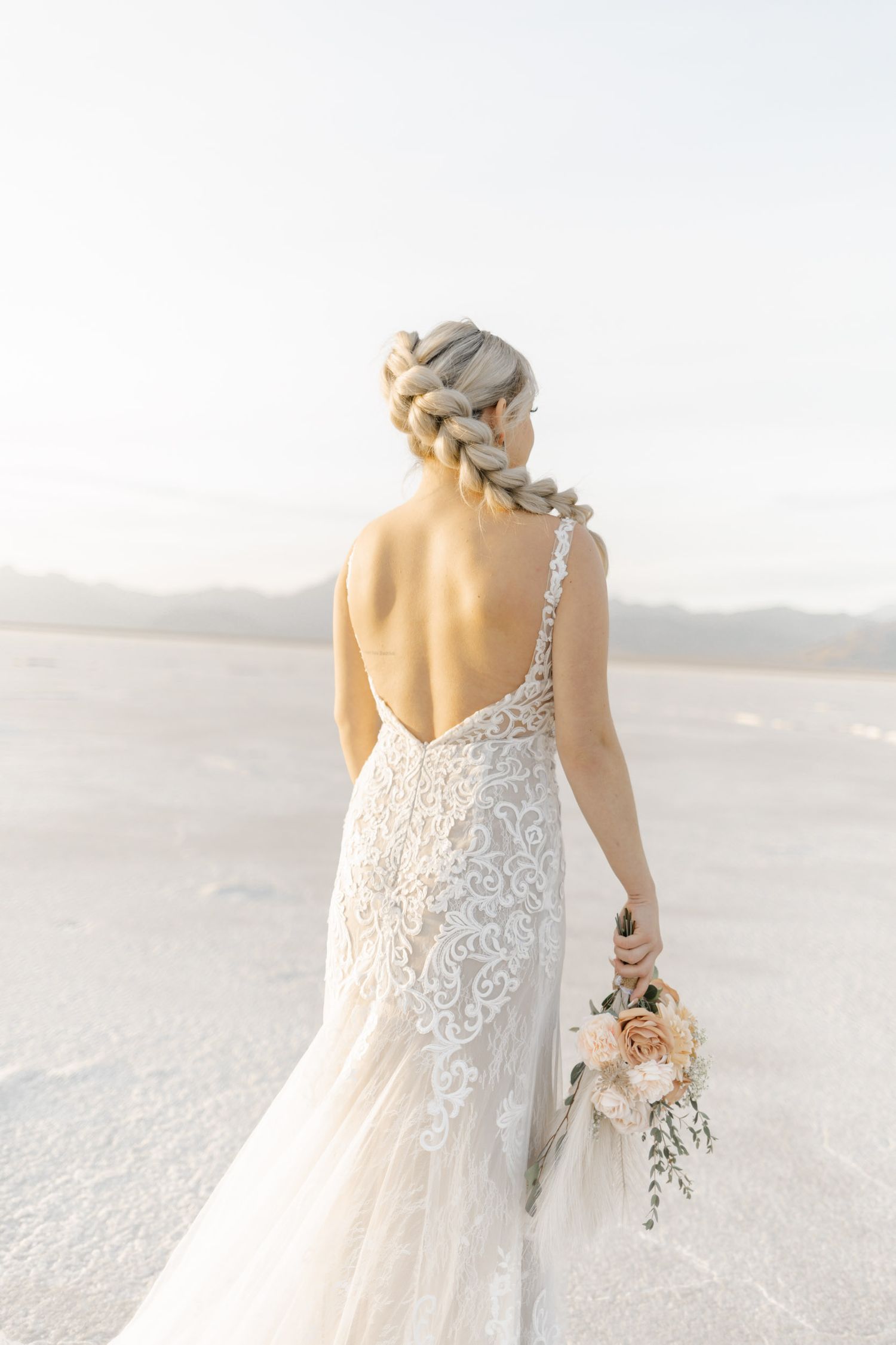 Elegant lace wedding dress shown from behind on a sandy beach with soft natural lighting and flowing fabric details.
