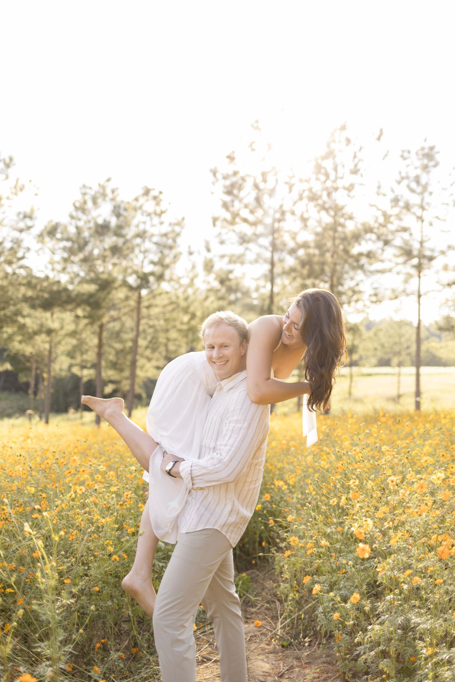 Playful couple in white clothing dancing and laughing in a field of yellow flowers at sunset during lifestyle portrait photoshoot.