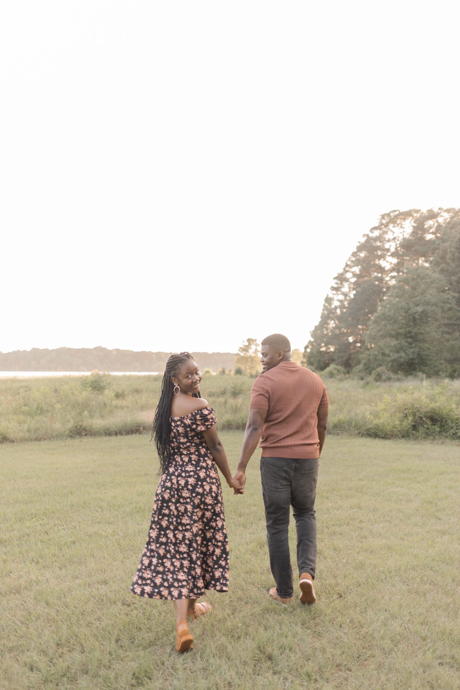 A Black couple holds hands walking through a green field near a lake at sunset, woman in floral dress looking back.