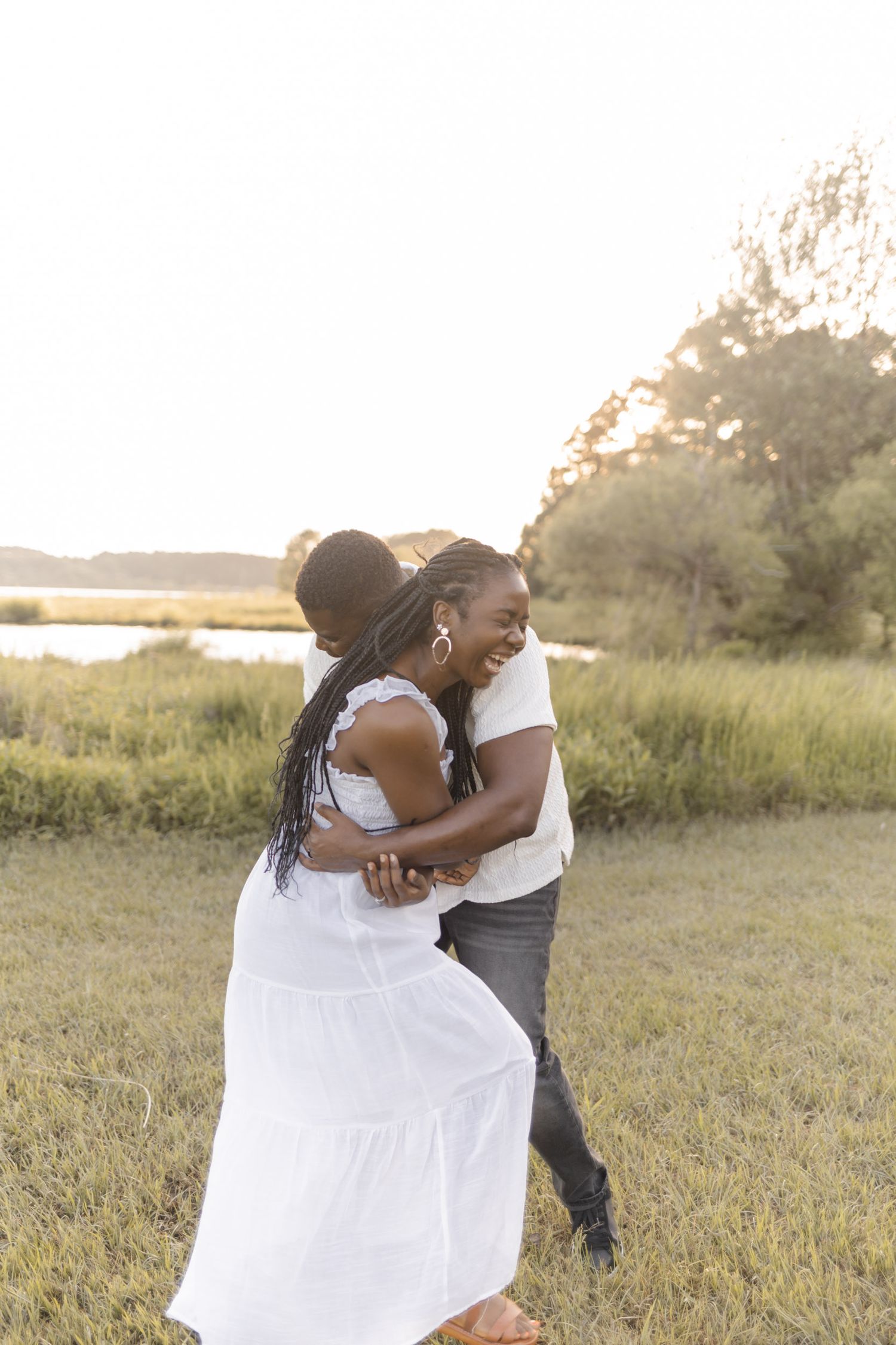 A joyful couple embraces and laughs together in a sunlit field near a marsh during golden hour.