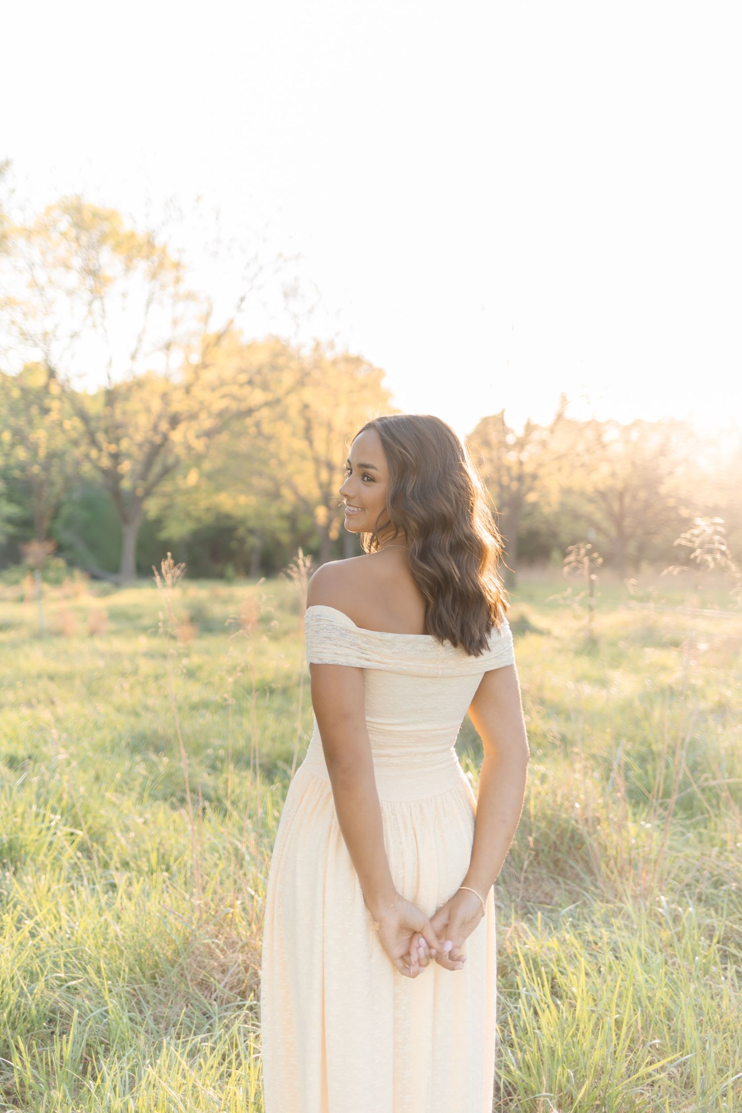 Young woman in off-shoulder cream gown stands in sunlit meadow, glancing back with flowing dark hair.