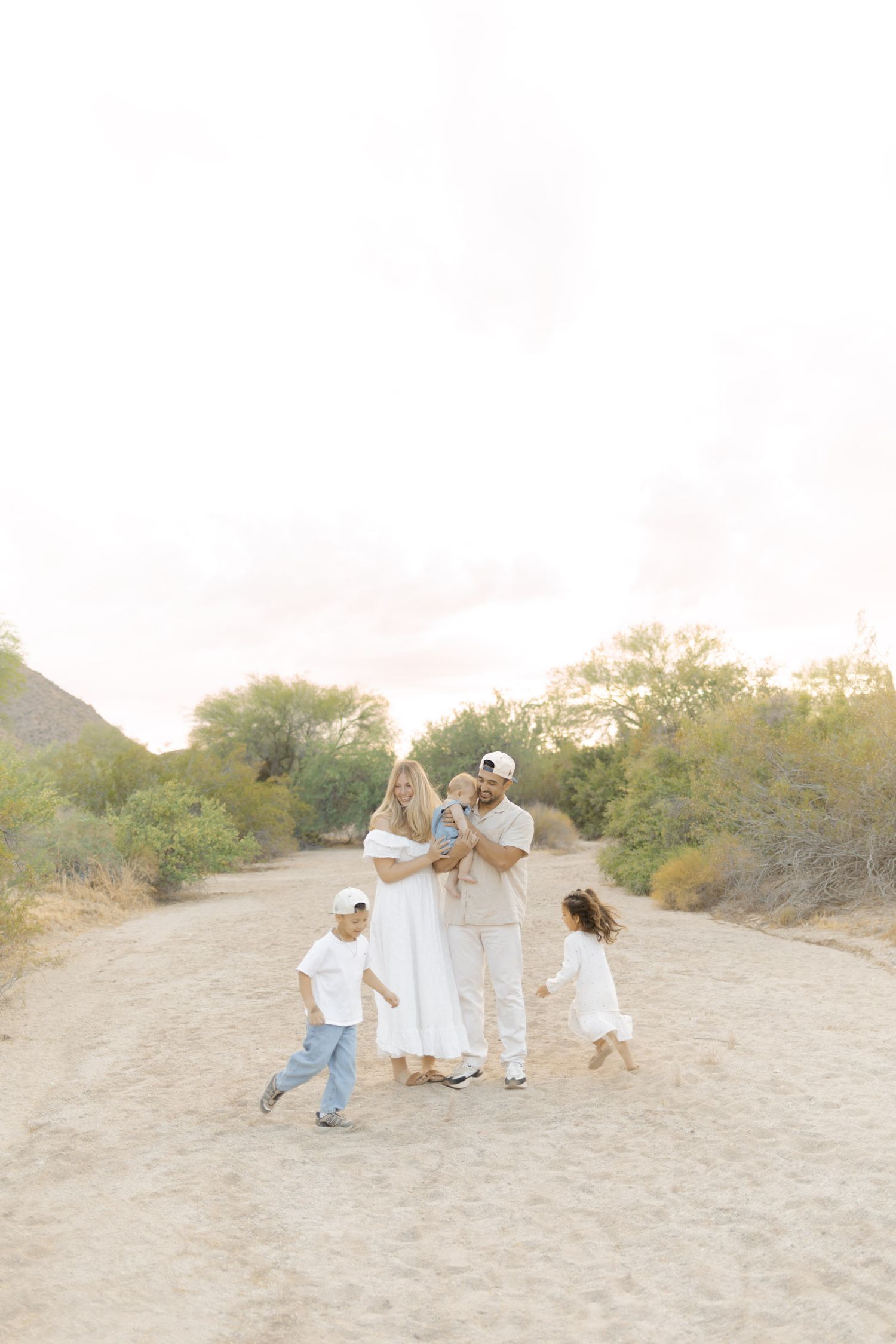 A young family of five dressed in white and beige poses together on a sandy desert trail at sunset.