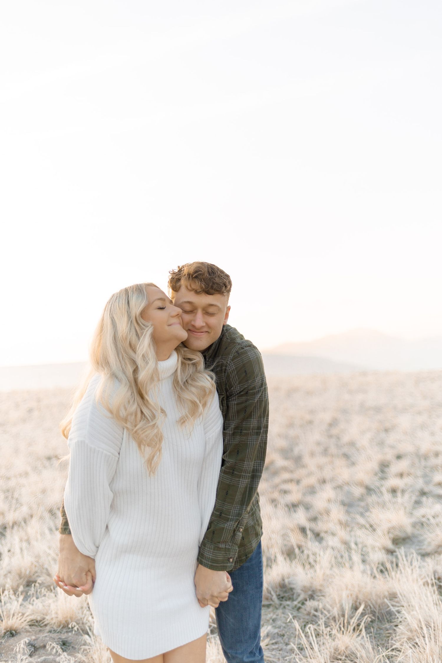 Couple sharing a tender kiss in a golden field at sunset, woman in white dress with long blonde hair.