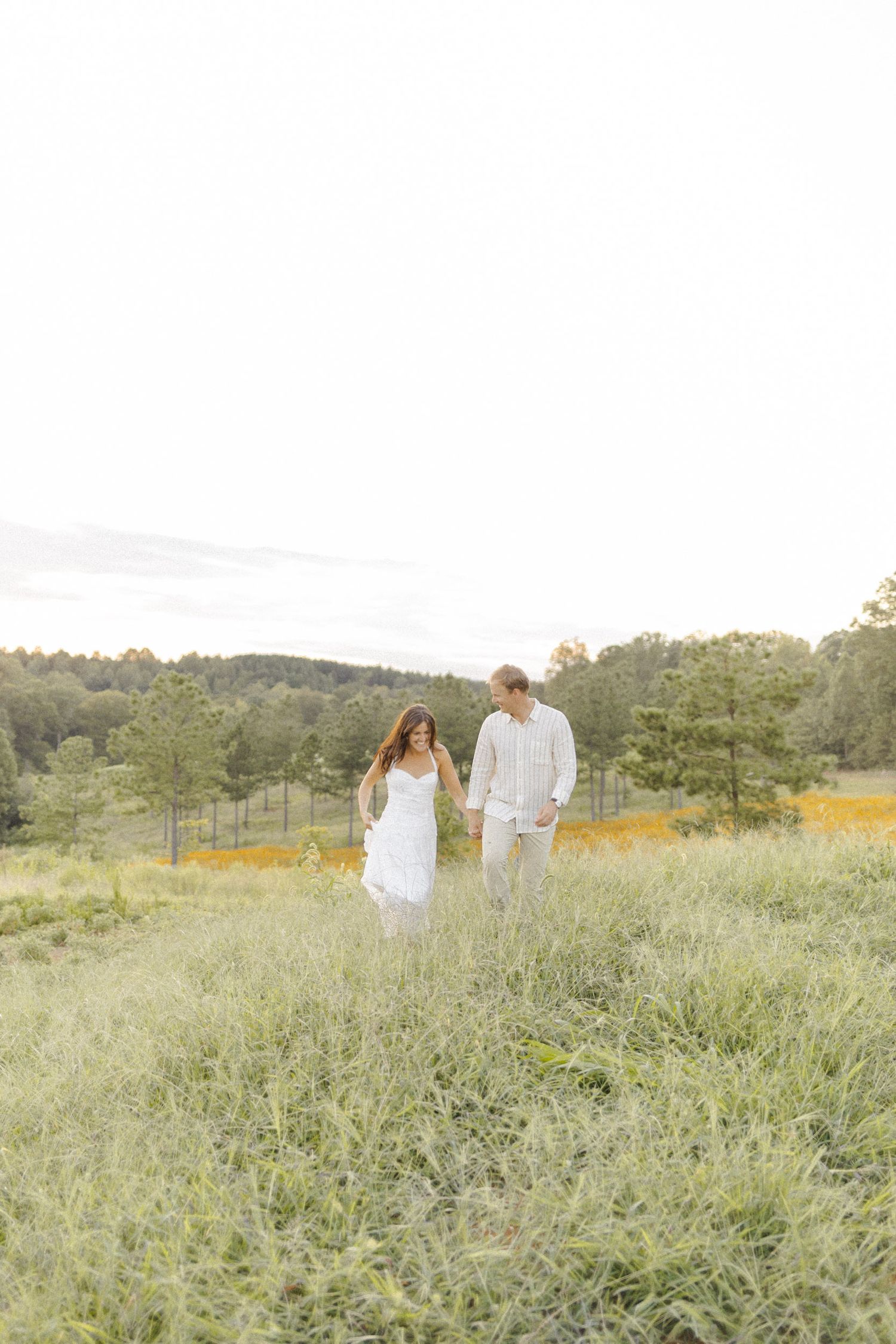 A couple in white outfits walks hand in hand through a lush green meadow with trees in the background.
