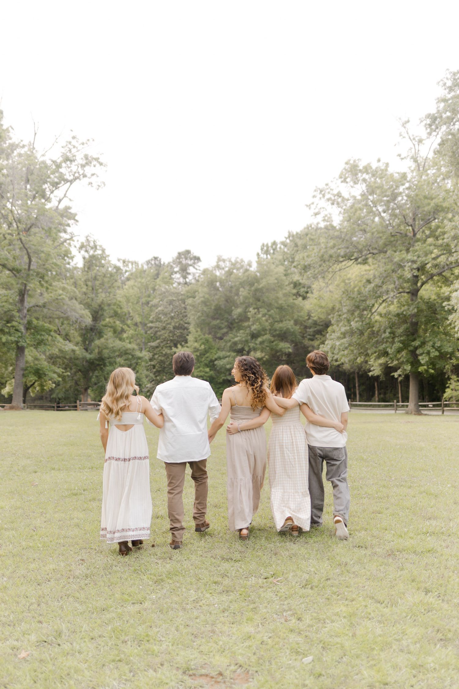 A group walks together arm-in-arm across a grassy field in coordinated light-colored outfits during photoshoot in Durham, NC.