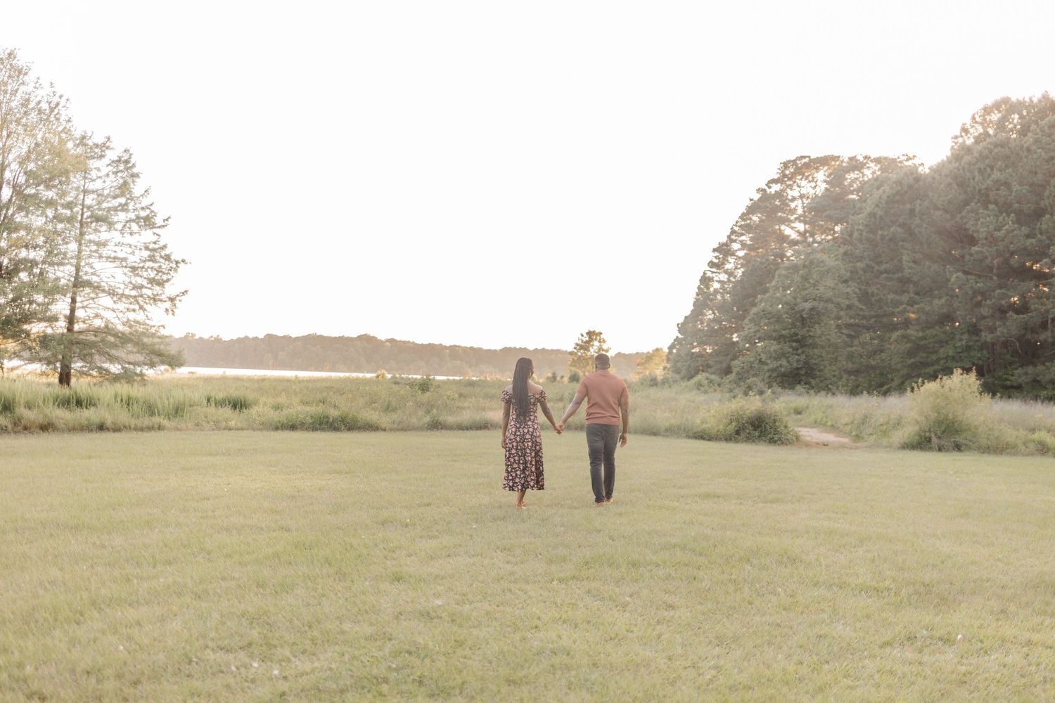 Couple holding hands walking through a lush green field at golden hour with trees and water in the background.