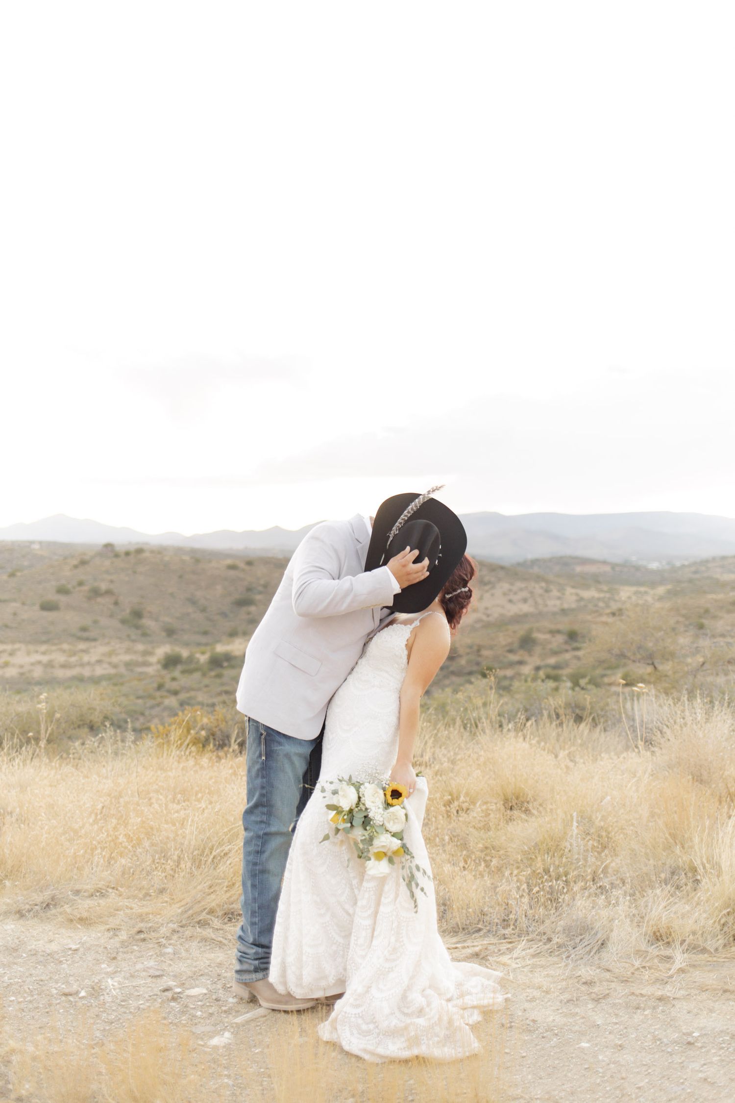 Bride and groom sharing a romantic kiss in a rustic desert landscape, bride holding a sunflower bouquet.