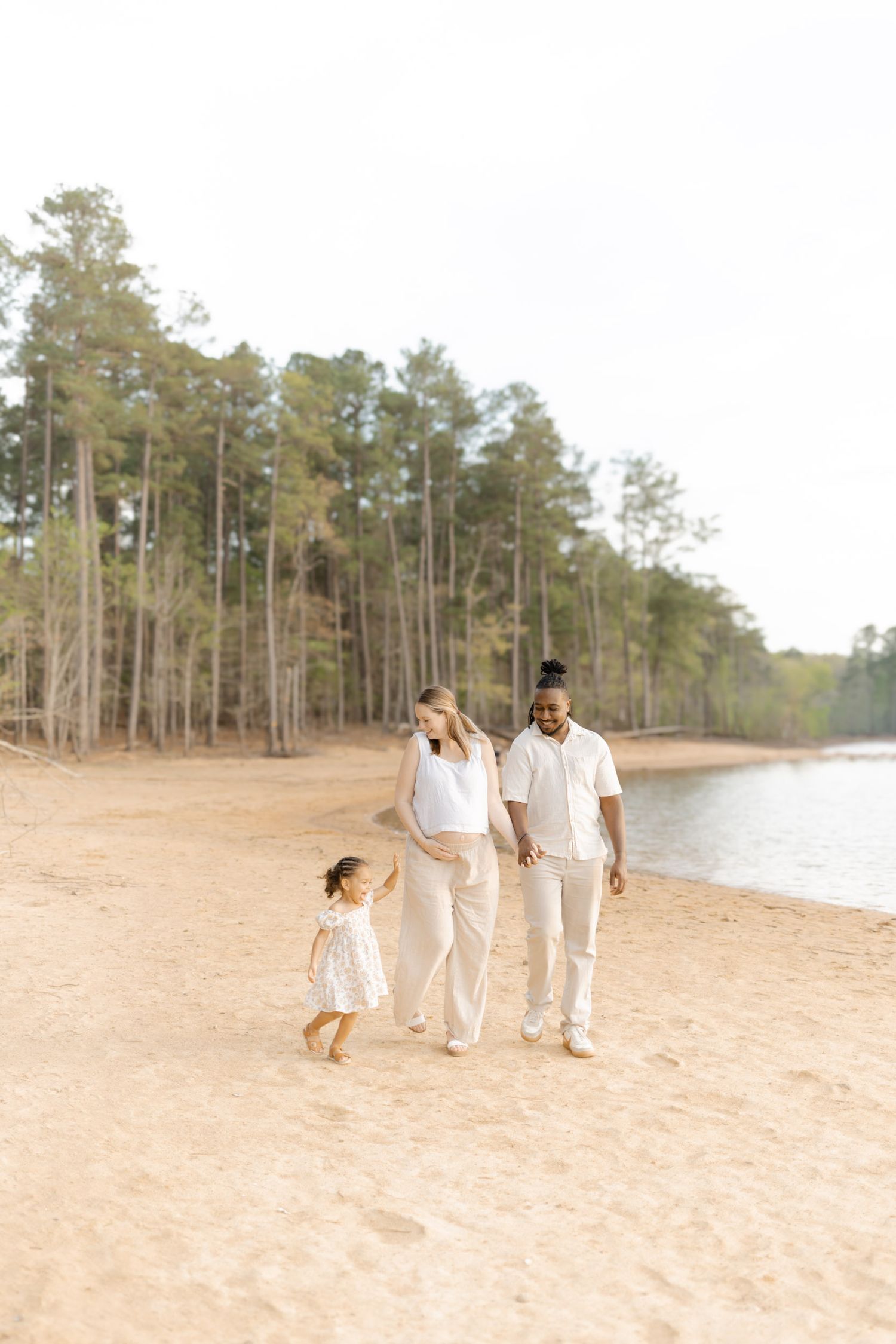A pregnant woman and her partner walk barefoot on a sandy beach with their young daughter.
