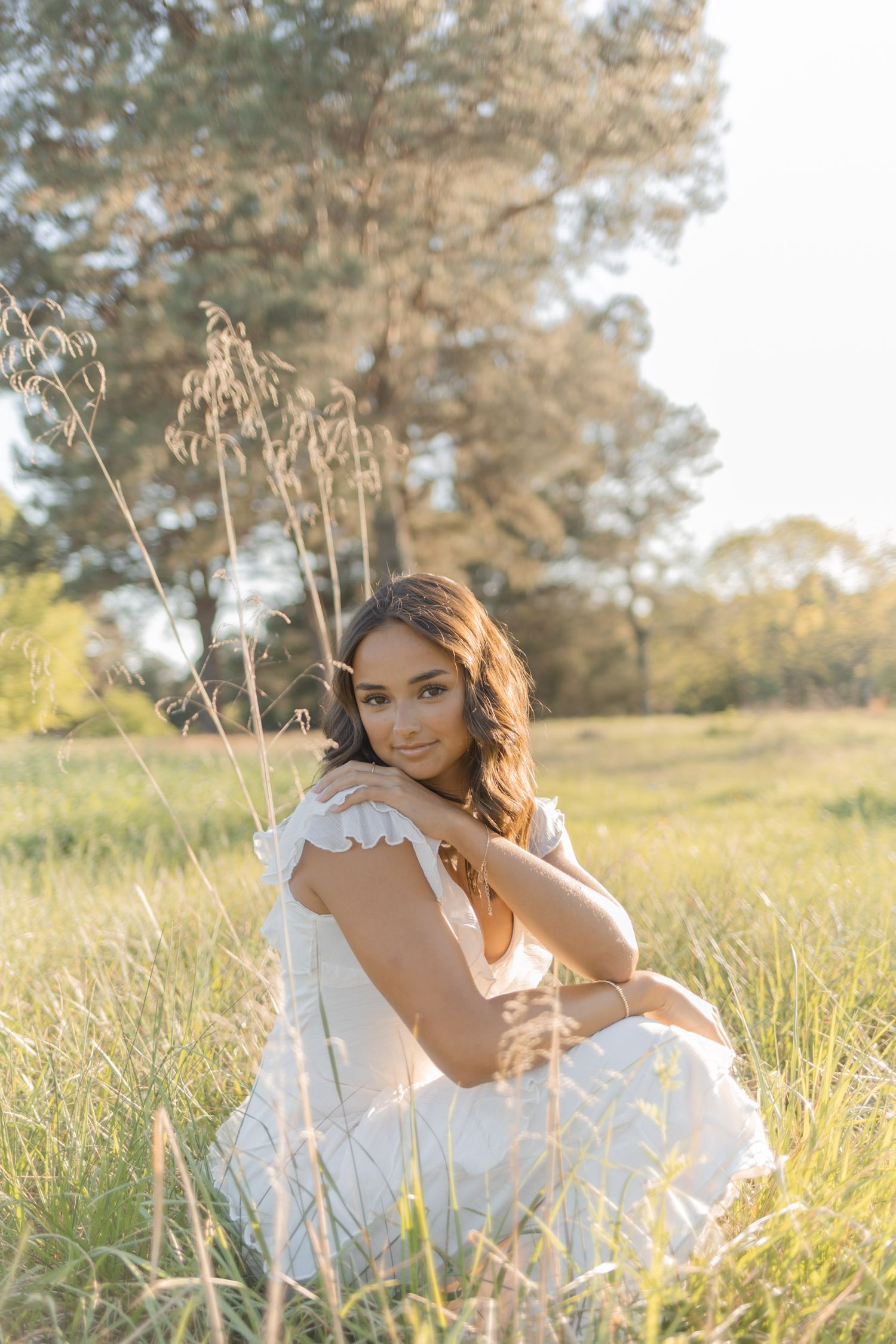 Young woman in white dress sitting in sunlit meadow with tall grass and trees, golden hour portrait photography.