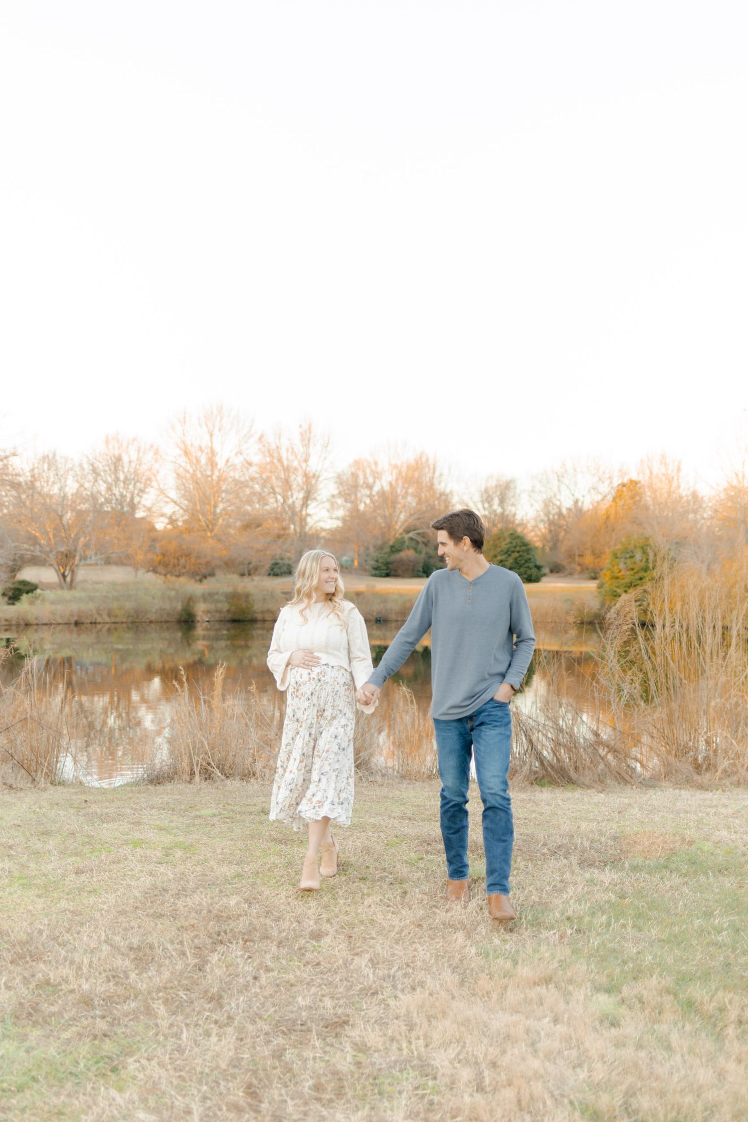 Couple holding hands walking in golden hour field near pond with bare trees in warm autumn light.