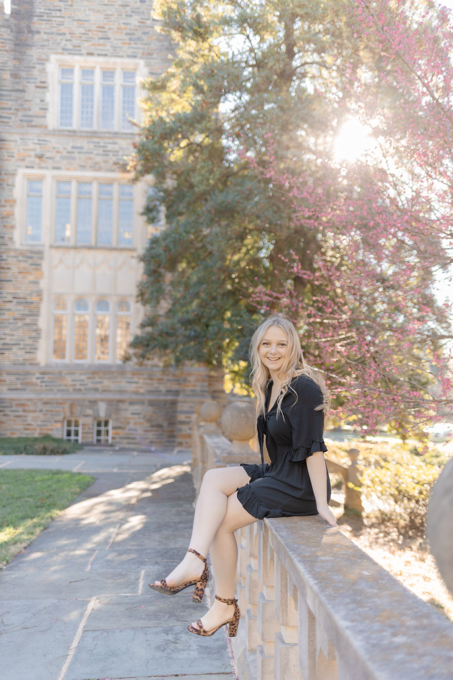 Young blonde woman in black dress and leopard heels sits on stone wall outside historic building with golden sunlight.